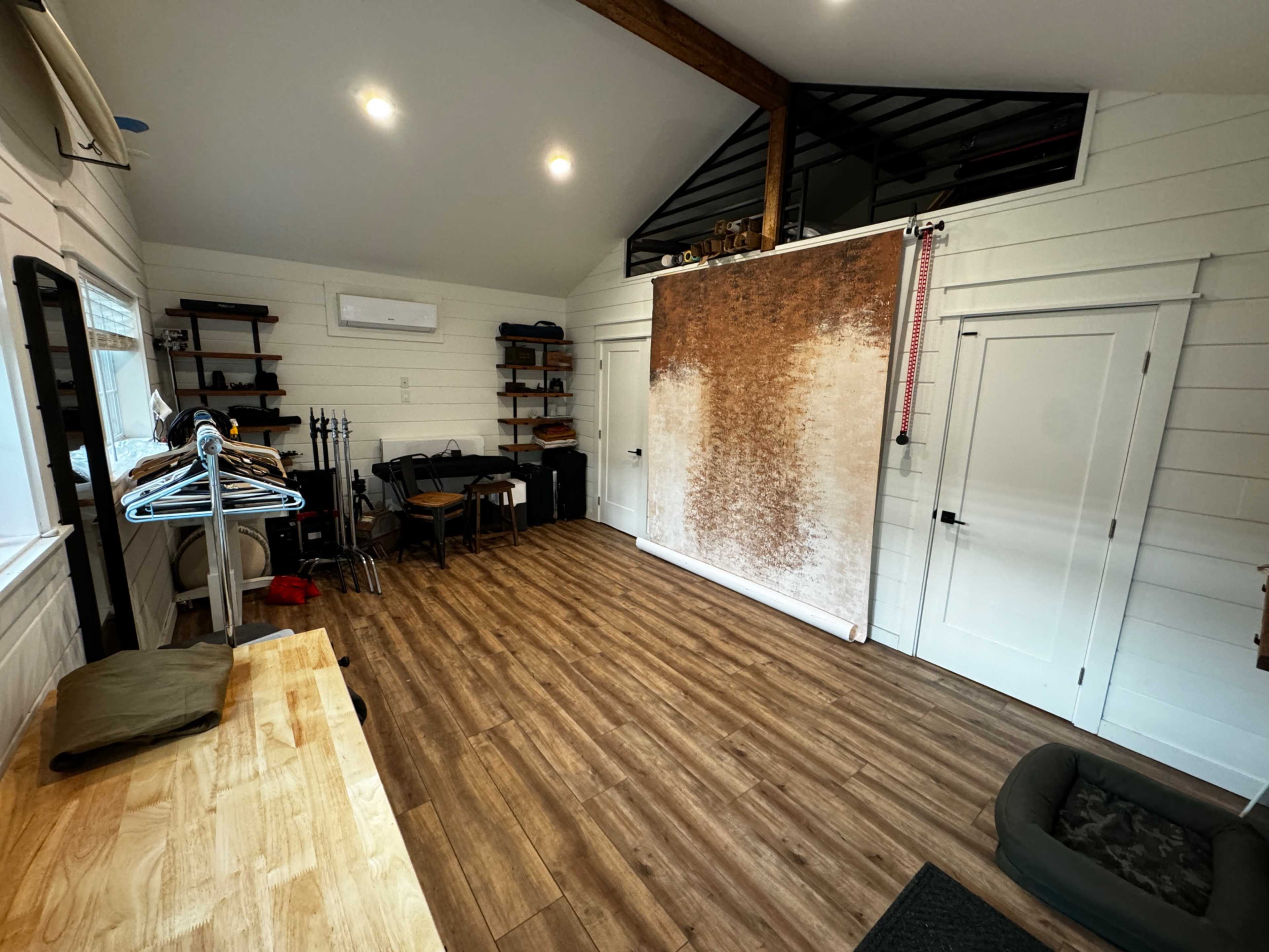The image shows a spacious, well-lit interior room with wooden flooring, a clothing rack, shelves, and a backdrop on the wall.