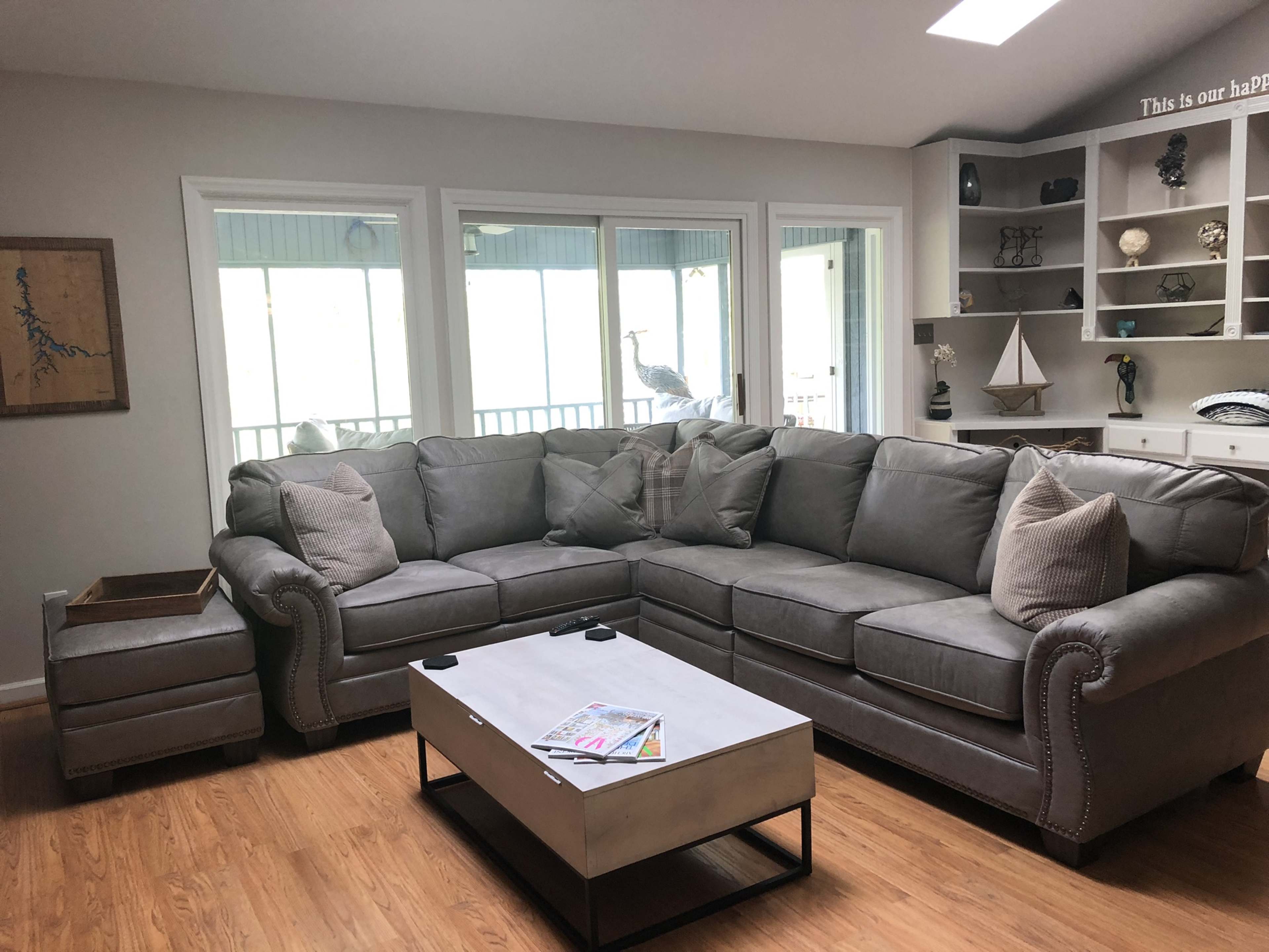 A gray sectional sofa with decorative pillows is arranged around a rectangular coffee table in a well-lit living room with large windows.