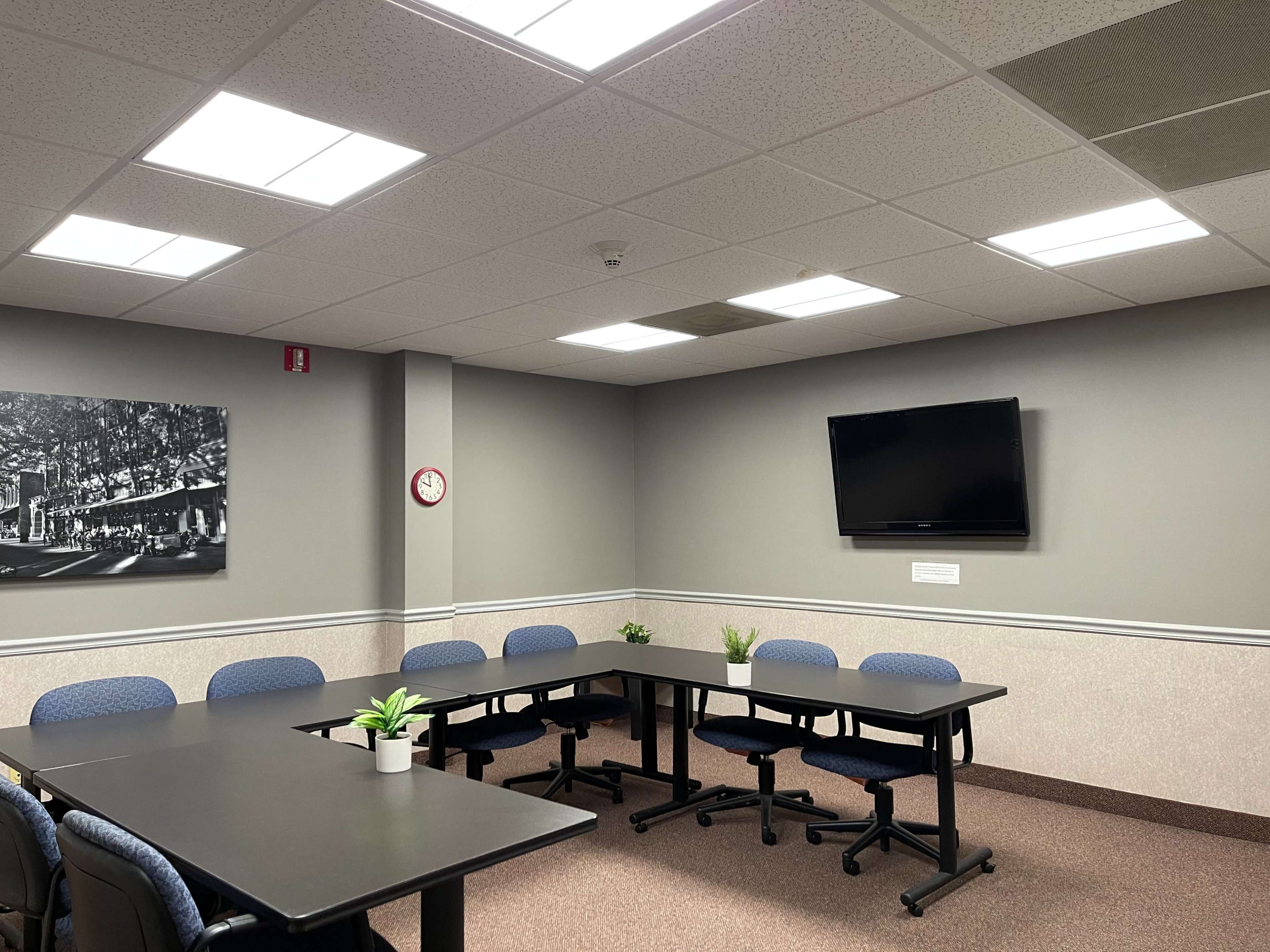 A conference room features a long table surrounded by blue chairs, with a mounted television on the wall and a clock beside a black and white photograph.