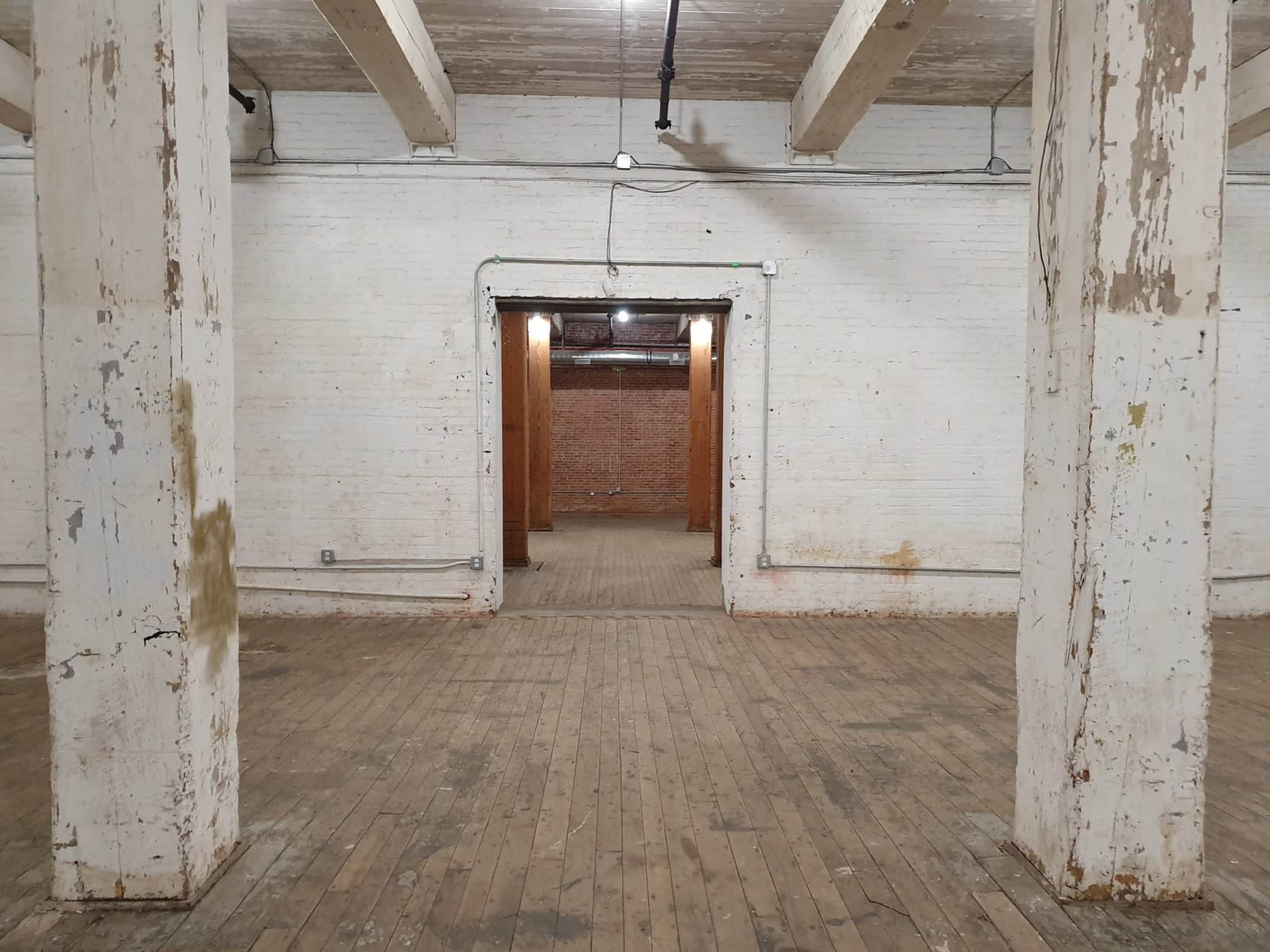 A spacious, empty room with wooden floors and large, white columns supporting the ceiling, leading to an arched doorway in the background.