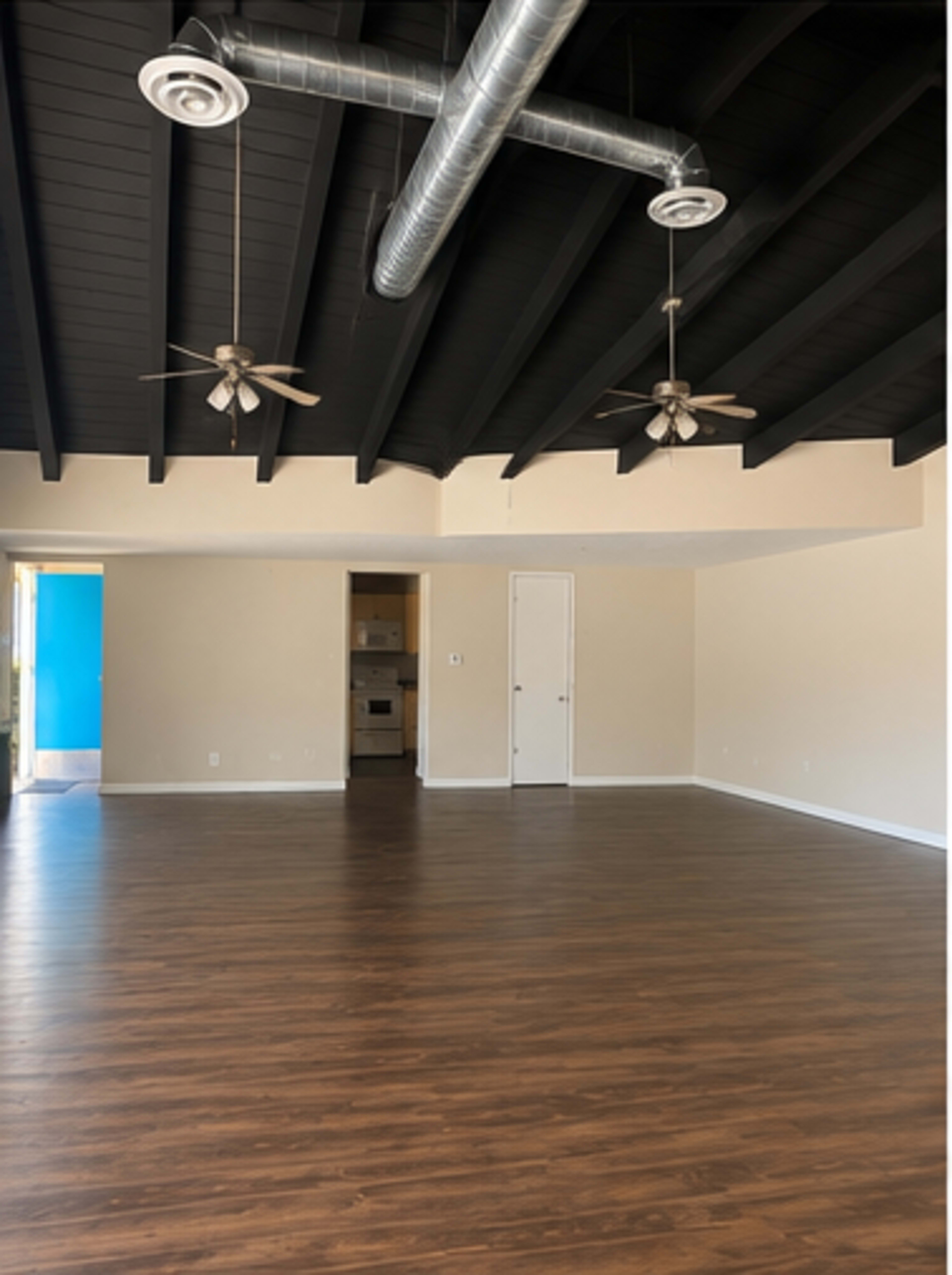 The image shows a spacious room with dark wood flooring, a vaulted ceiling featuring exposed beams, and two ceiling fans.