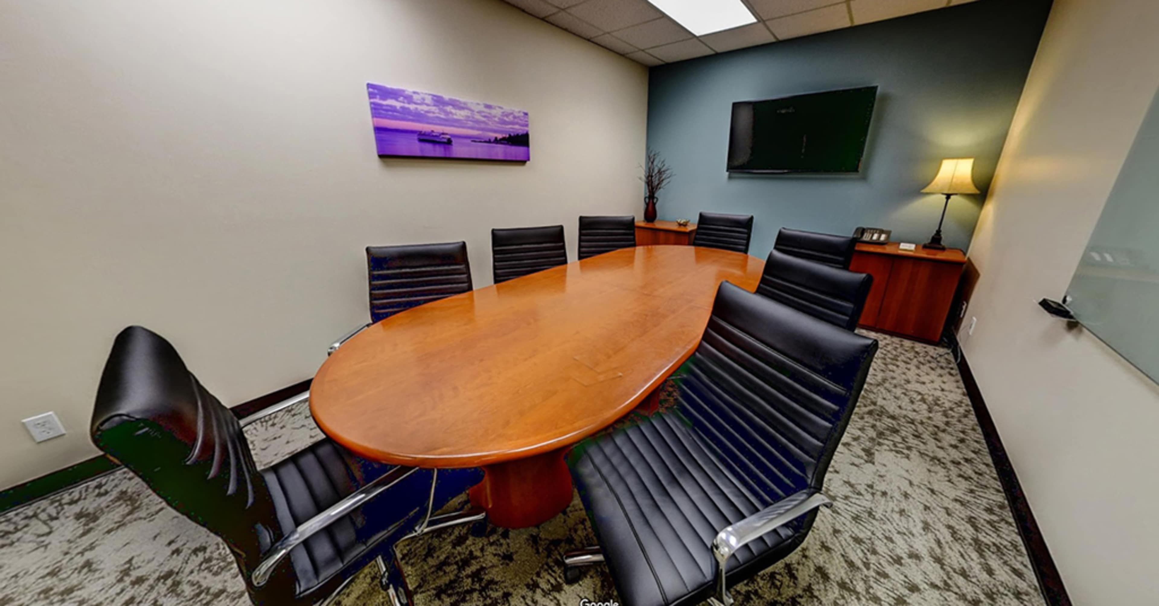 A conference room features an oval wooden table surrounded by black leather chairs, with a television on the wall and a lamp in the corner.