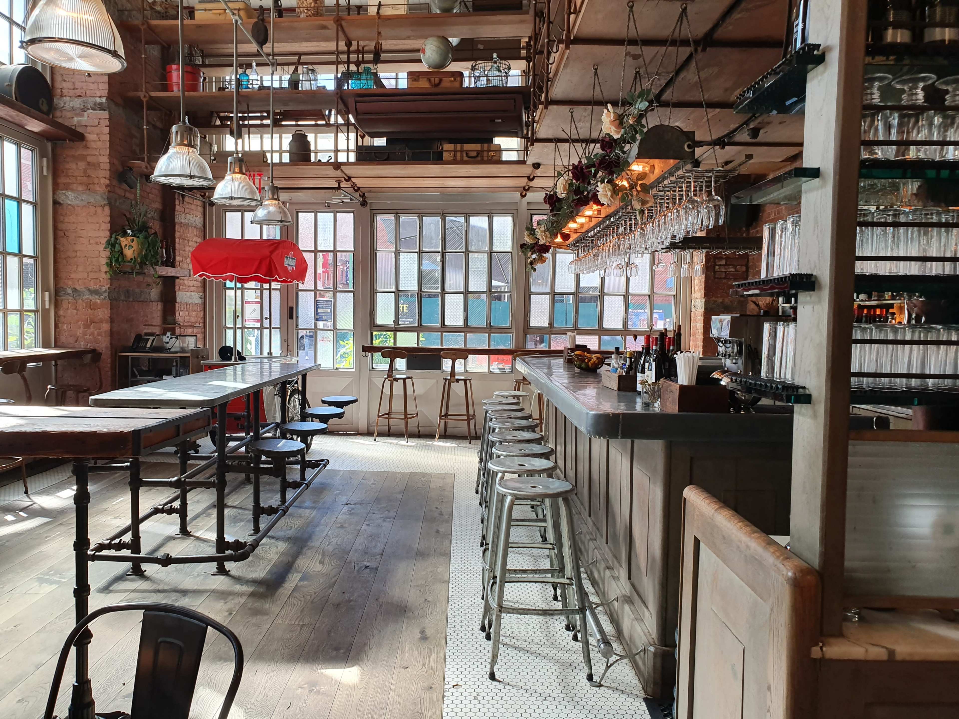 An industrial-style restaurant interior with wooden floors, a bar lined with stools, and large windows allowing natural light to flow in.