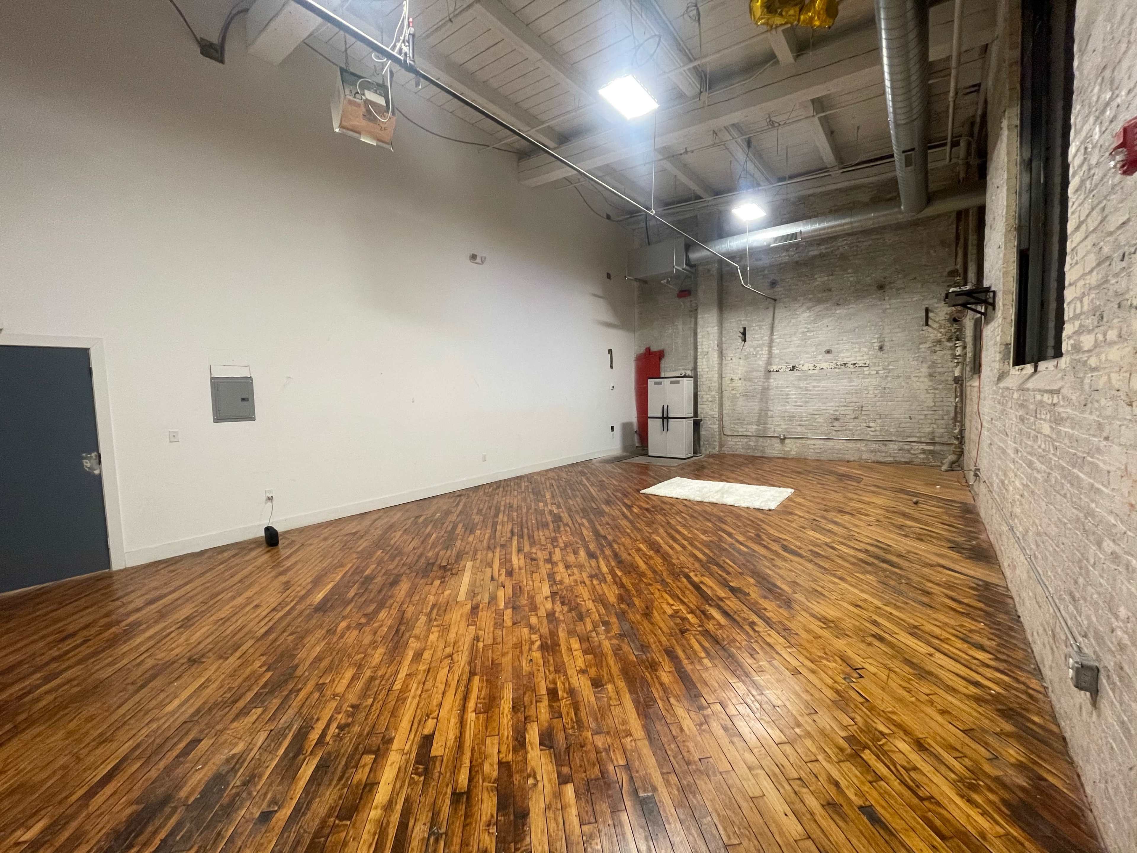 The image shows a spacious, empty room with hardwood flooring and exposed brick walls, featuring a white patch on the floor and a gray door on one side.