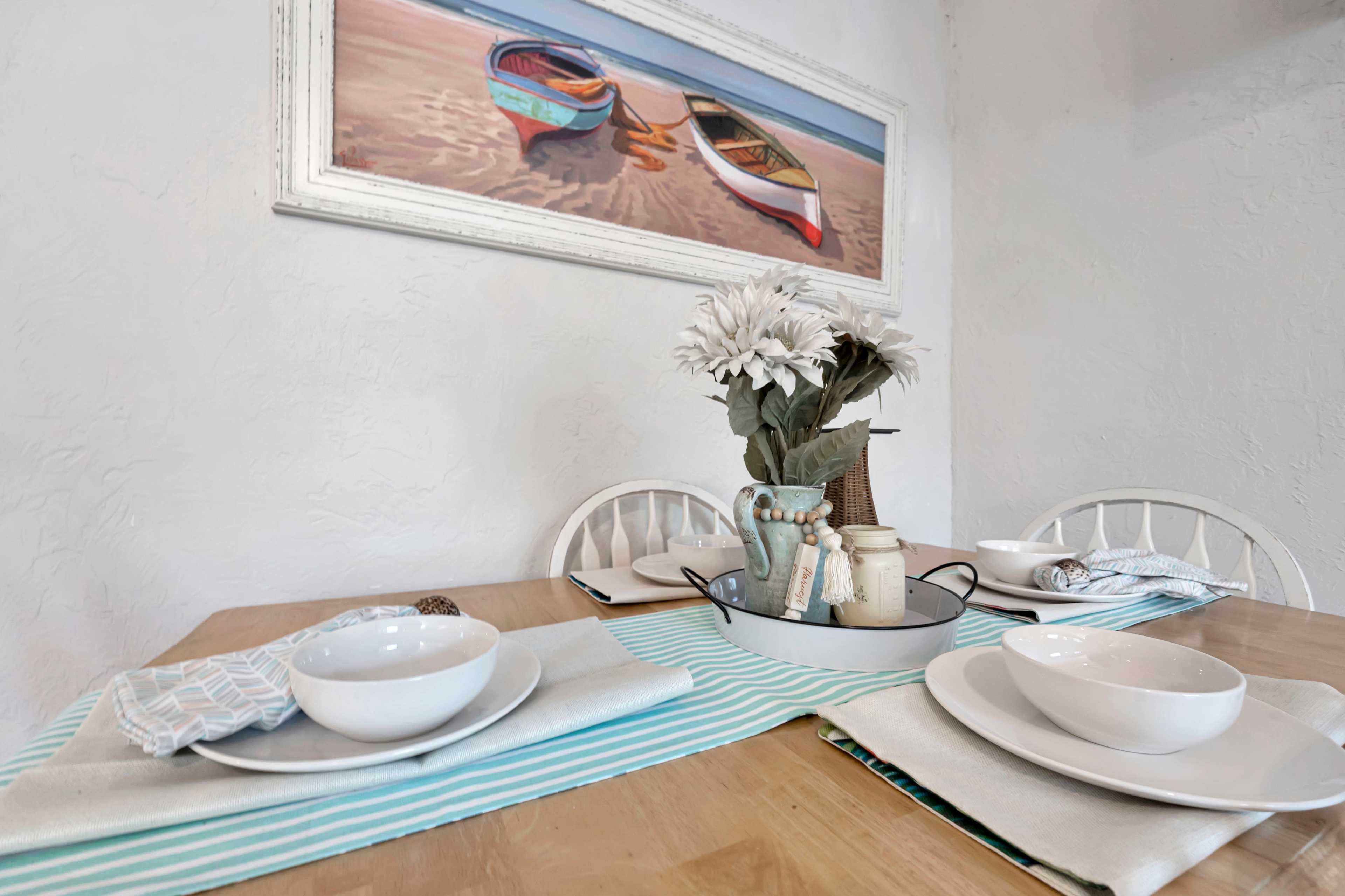 A dining table is set with white bowls, placemats, and a centerpiece of flowers, under a coastal-themed painting of boats.