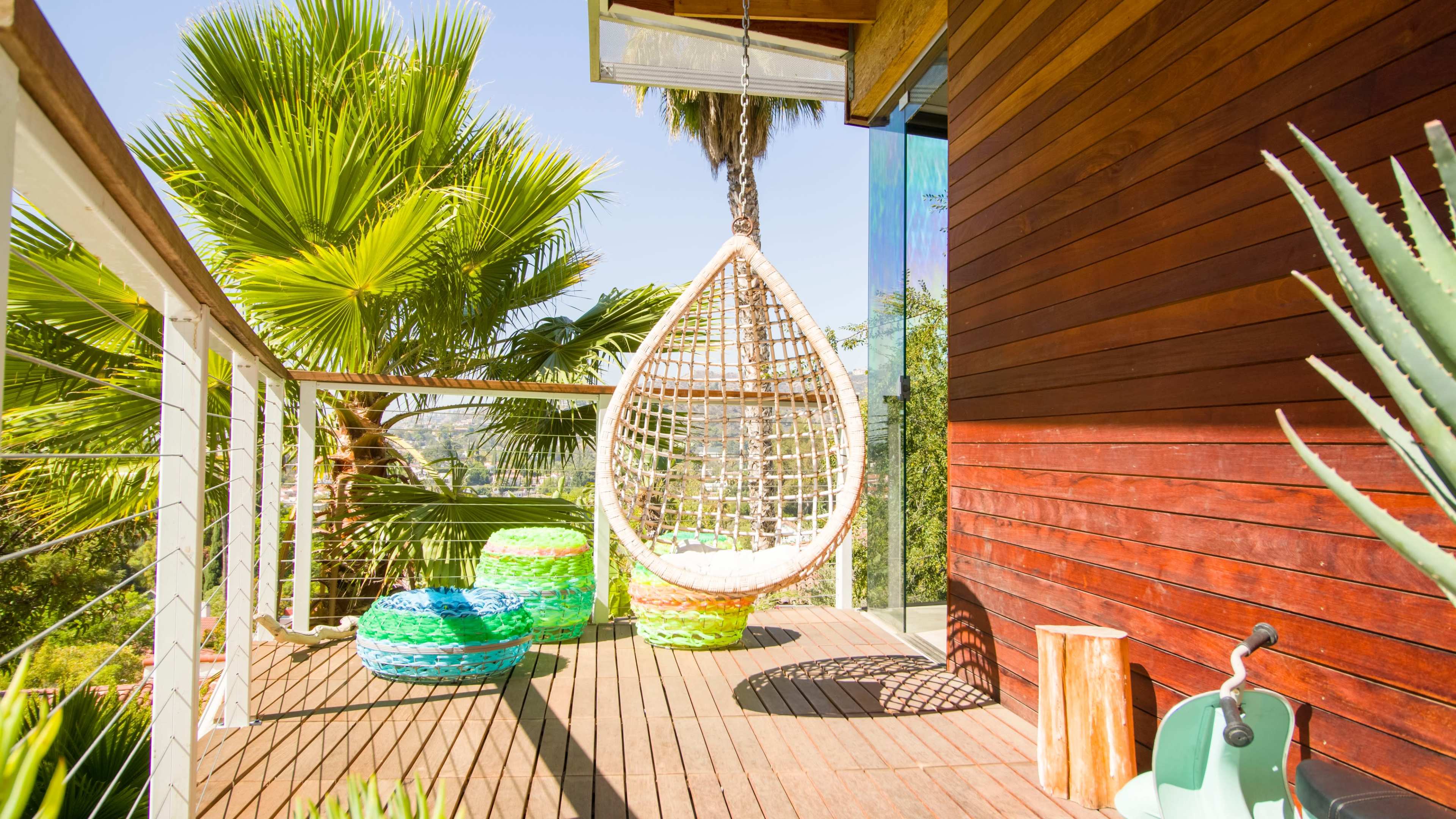 A hanging wicker chair is positioned on a wooden deck beside colorful poufs and surrounded by lush palm trees.
