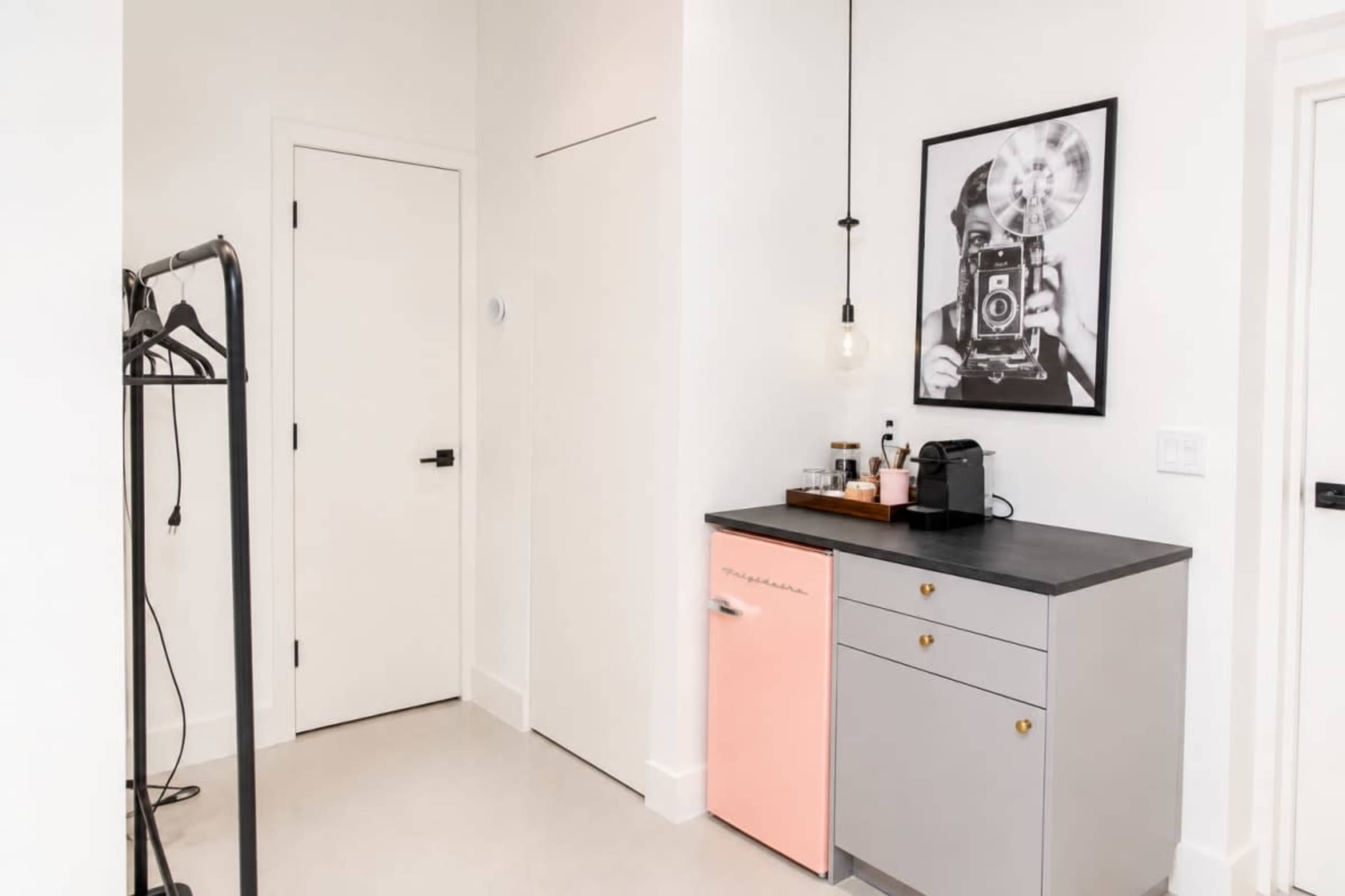 The image shows a minimalist entryway featuring a small pink refrigerator, a coffee maker, and a framed photograph on the wall, complemented by a coat rack.