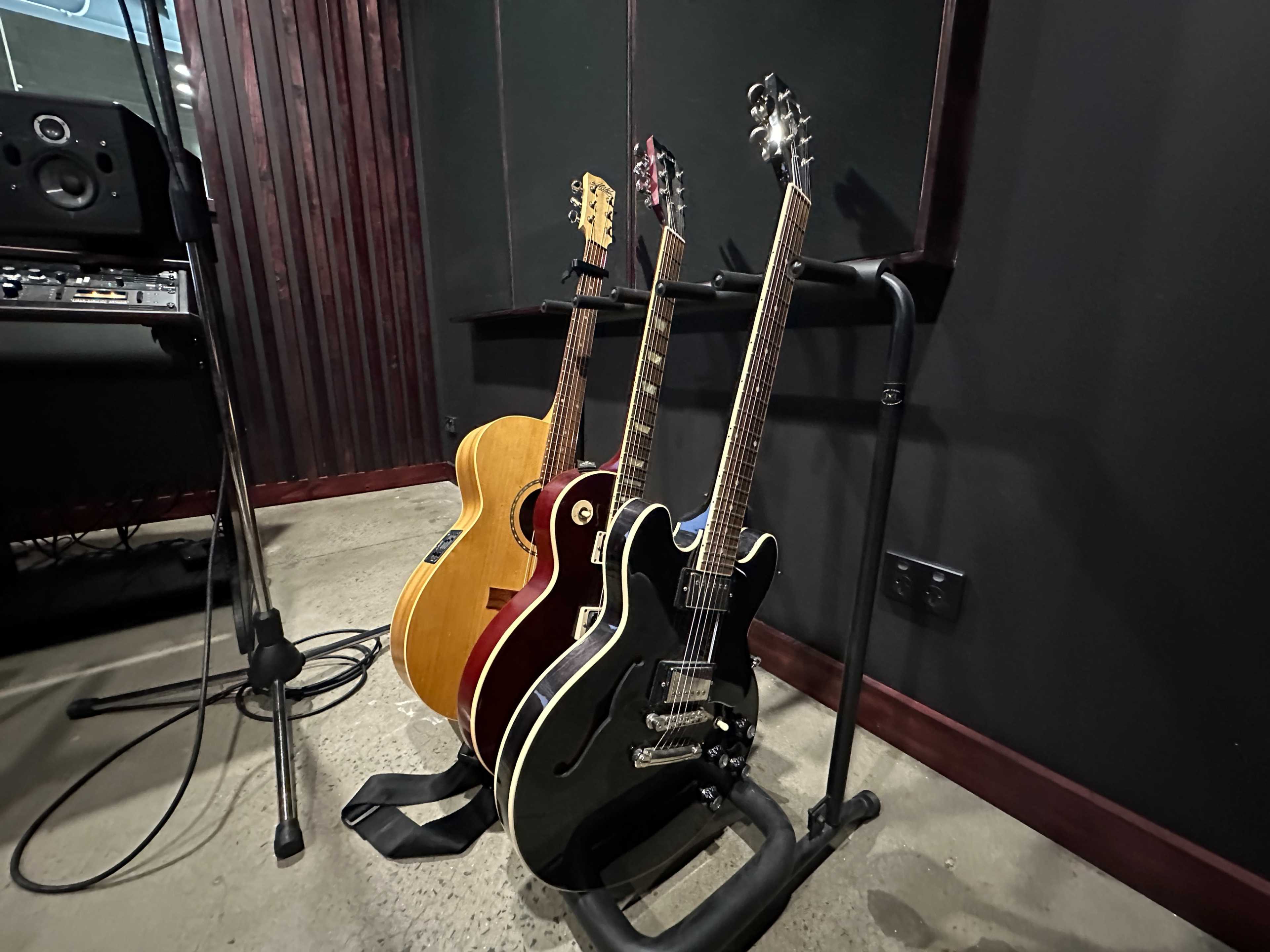 Three guitars, including an acoustic and two electric models, lean against a music stand in a recording studio.