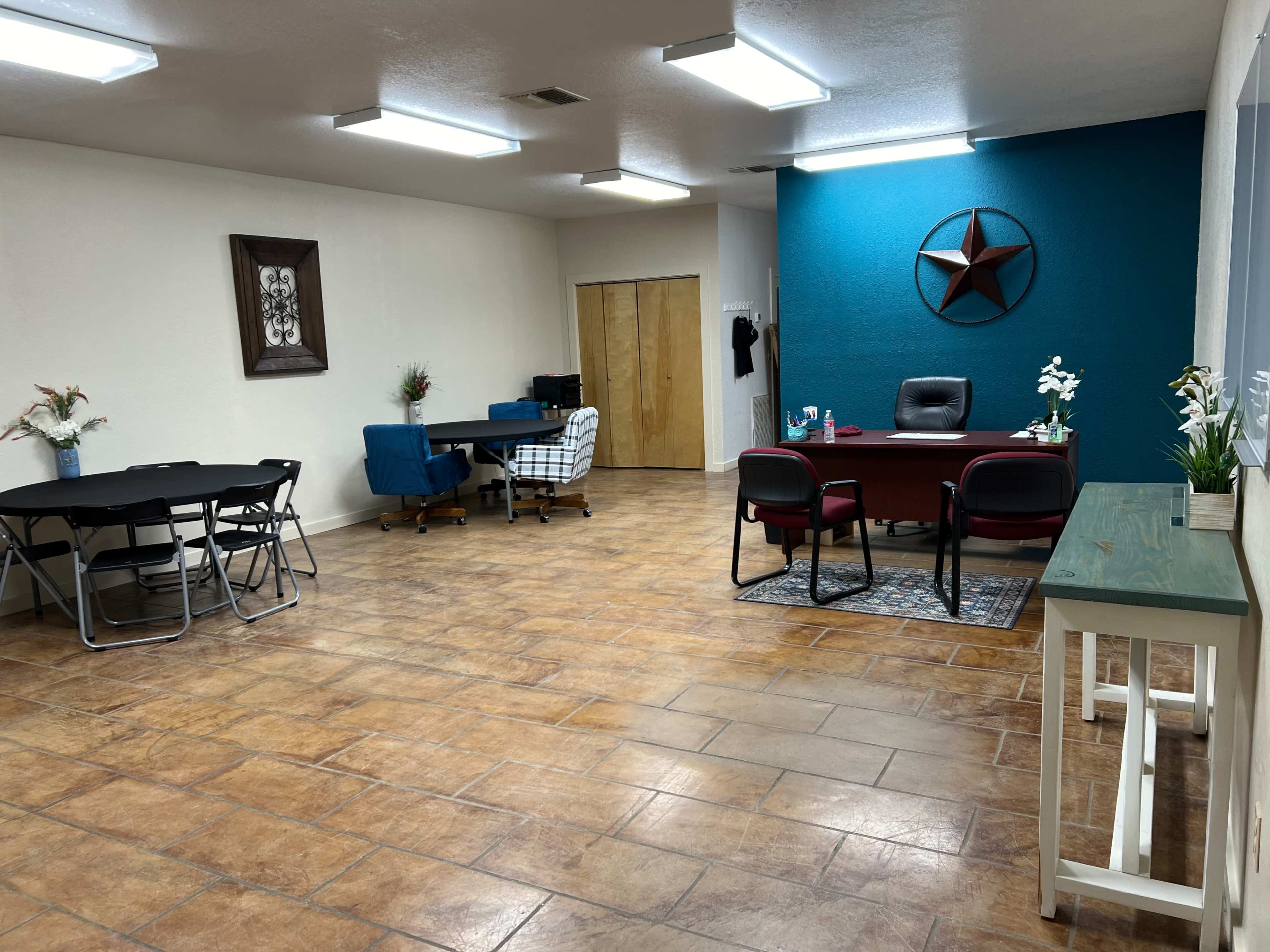The image shows an empty room with tiled flooring, featuring a desk with a chair, a blue wall with a star decoration, a folding table, and a couple of seating areas.