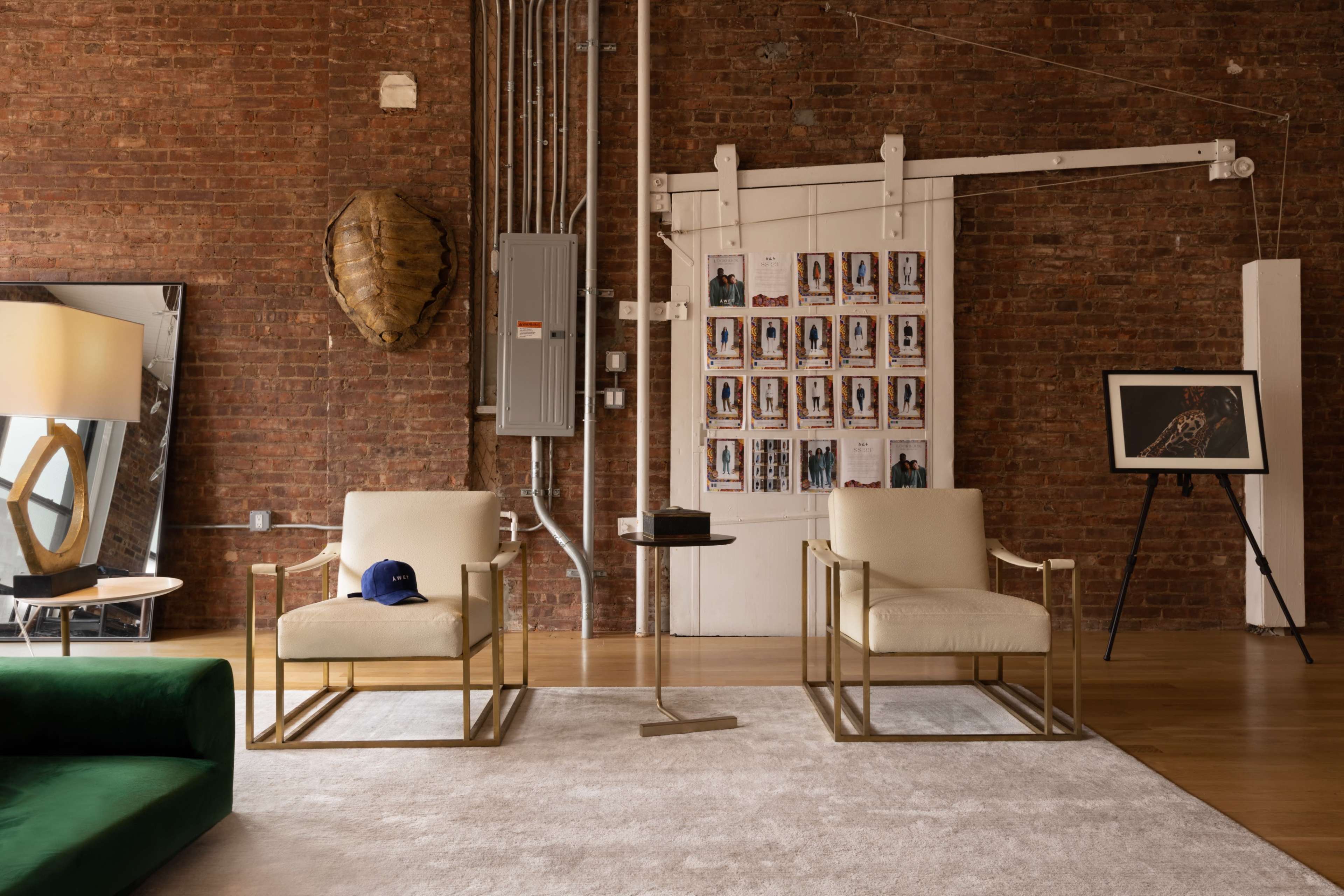 The scene features a cozy living area with two gold-framed chairs, a small side table, and a large mirror against a brick wall, complemented by artwork and a baseball cap on one of the chairs.