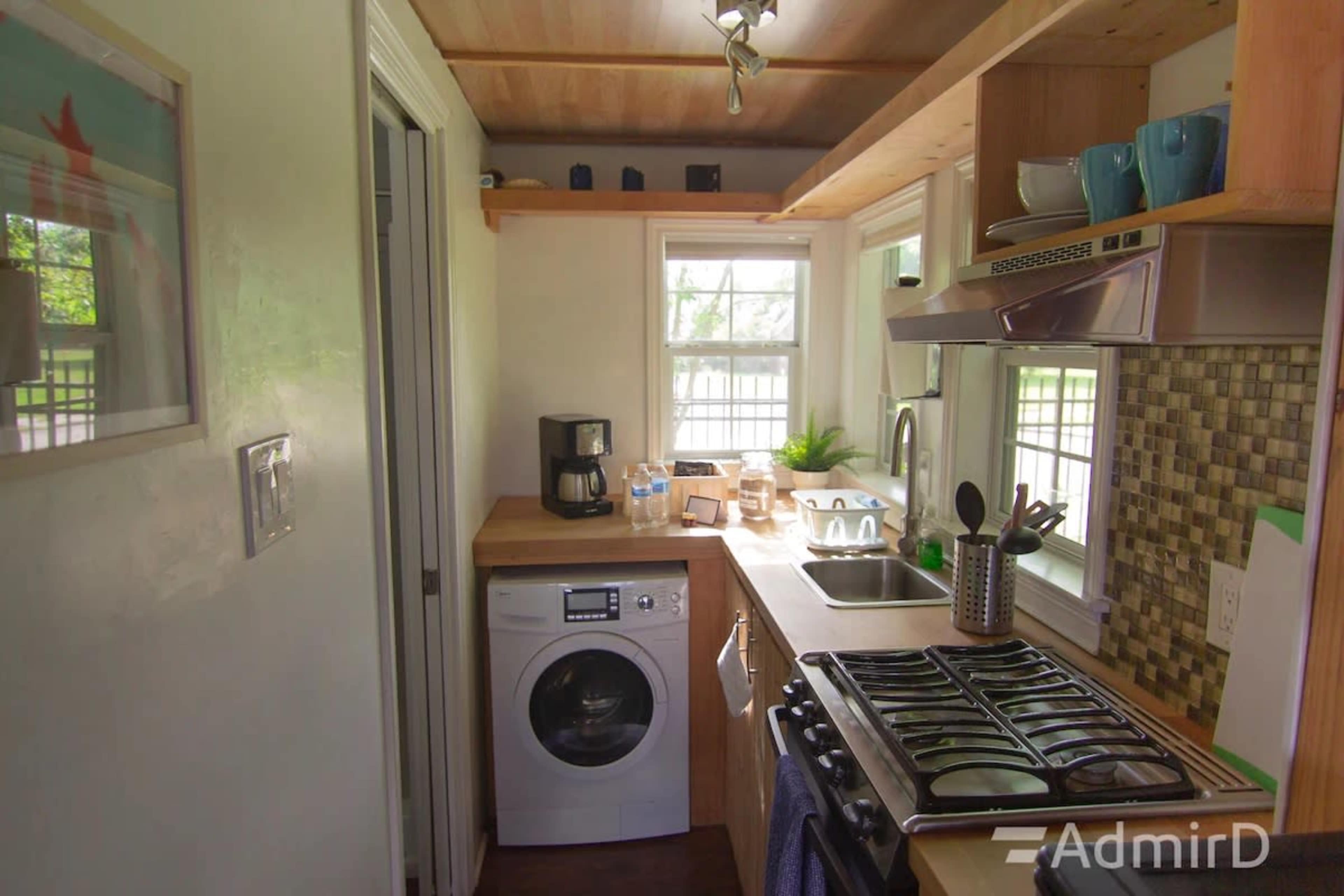 The image shows a compact kitchen area with a washing machine, a sink, a stove, and open shelving displaying kitchenware.