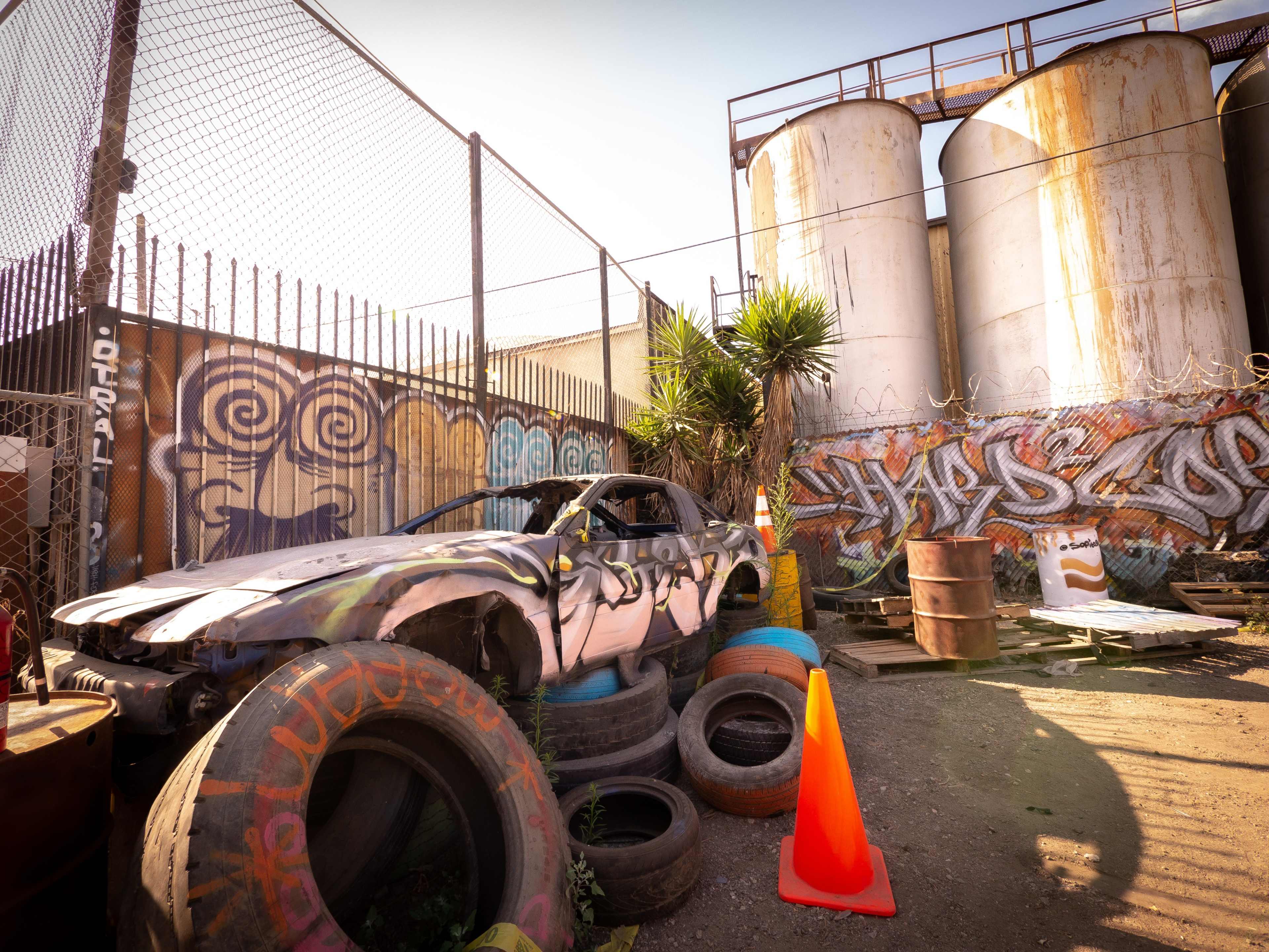 An abandoned car with graffiti is surrounded by old tires and industrial storage tanks in a fenced area.