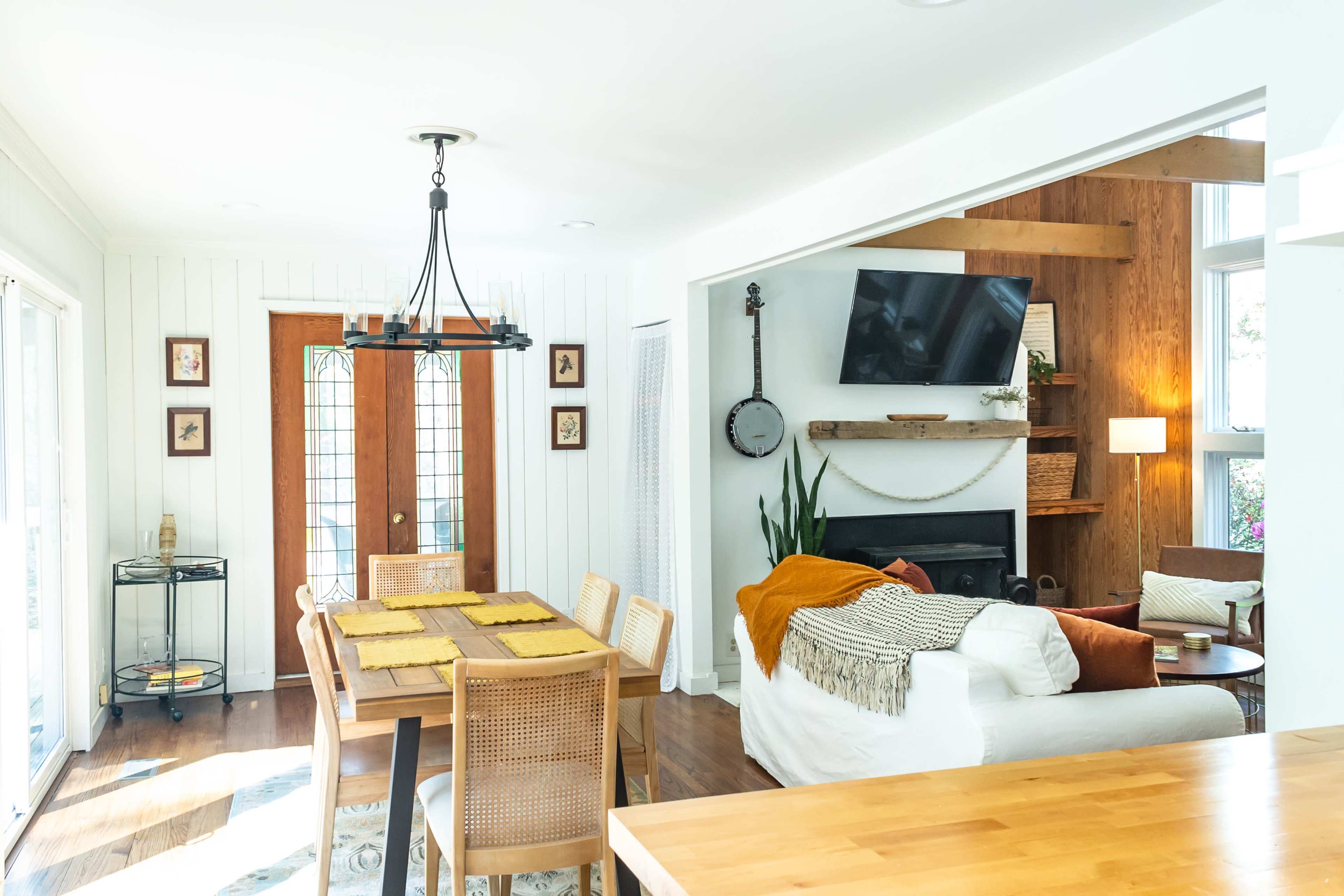 A bright, modern living space featuring a dining area with a wooden table and chairs, a cozy sitting area with a television, and warm wood accents.