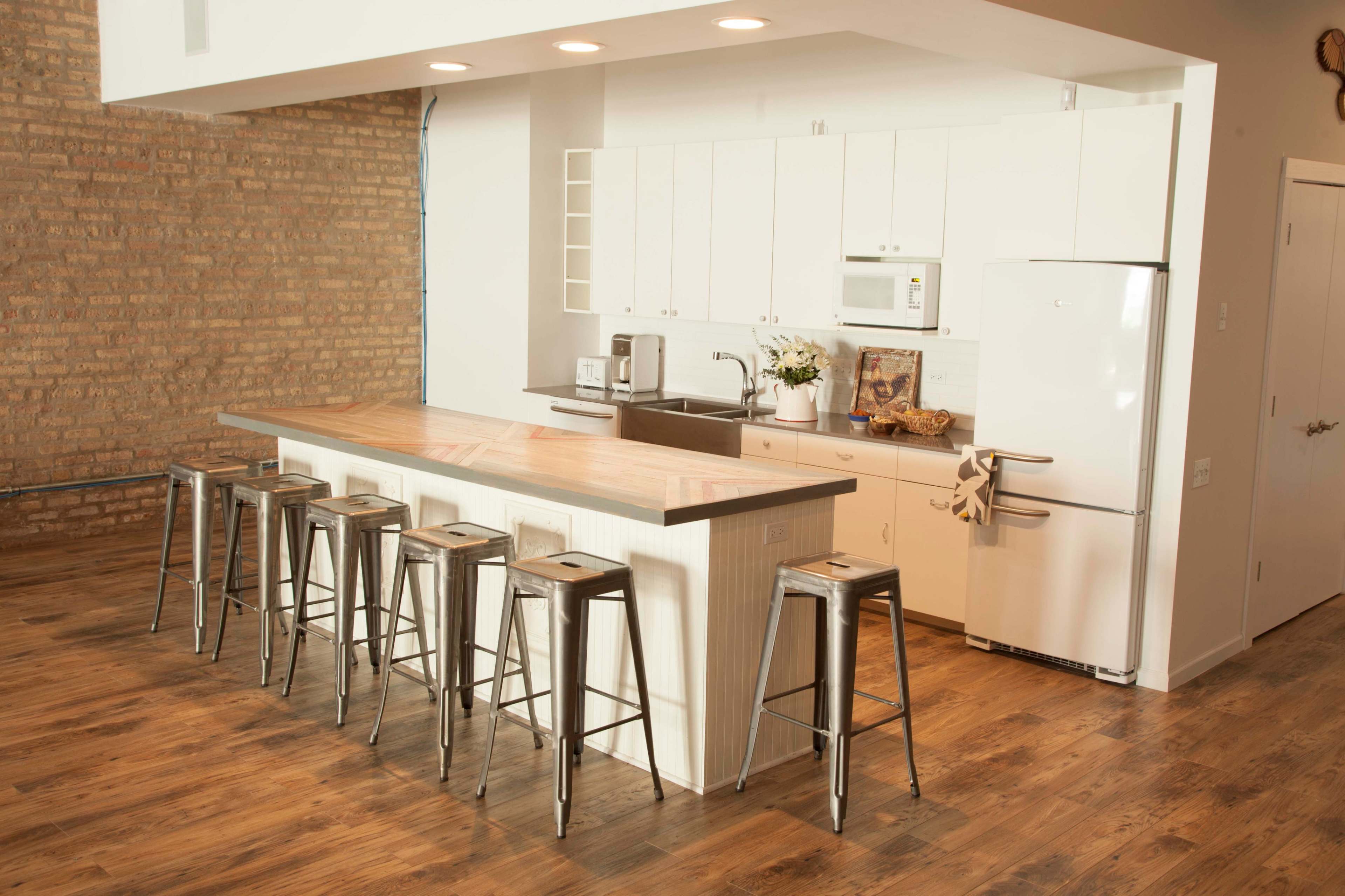 The image shows a modern kitchen featuring a long wooden island with metal bar stools, white cabinetry, a refrigerator, and exposed brick walls.
