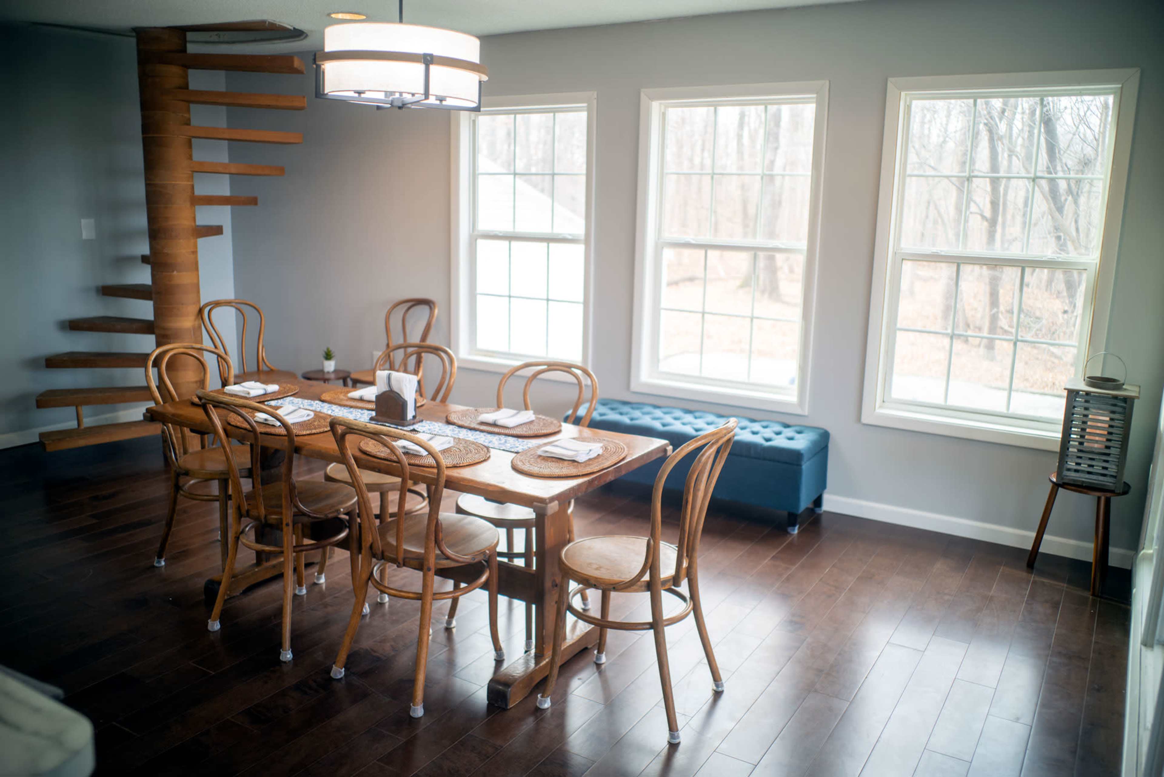 A wooden dining table with eight chairs is positioned in a well-lit room featuring large windows and a spiral staircase.