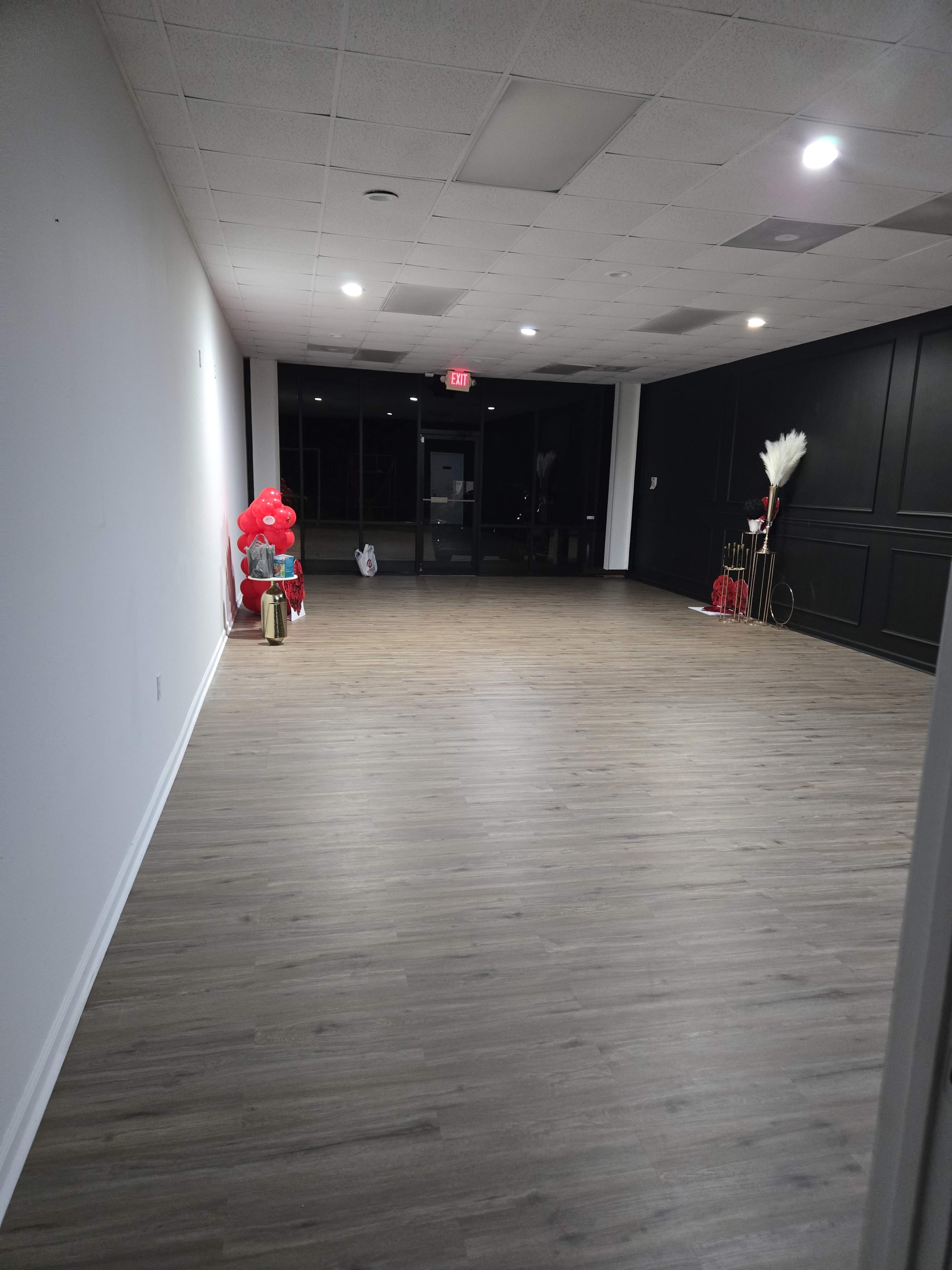 The image shows a spacious, empty room with wooden flooring and a wall lined with decorative items on one side.