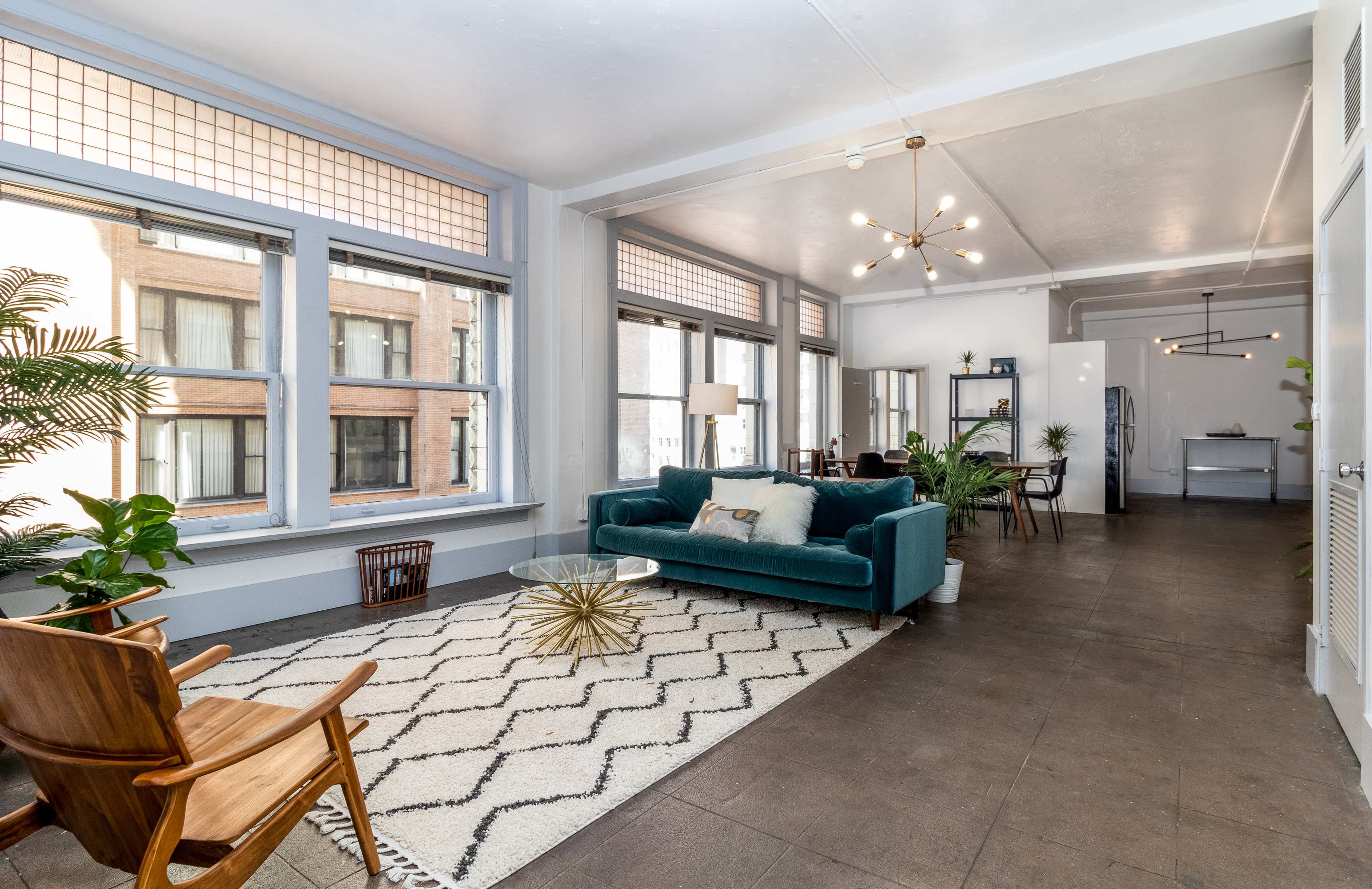 The image shows a bright, spacious living area featuring a teal sofa, a patterned rug, and large windows that allow natural light to fill the room.
