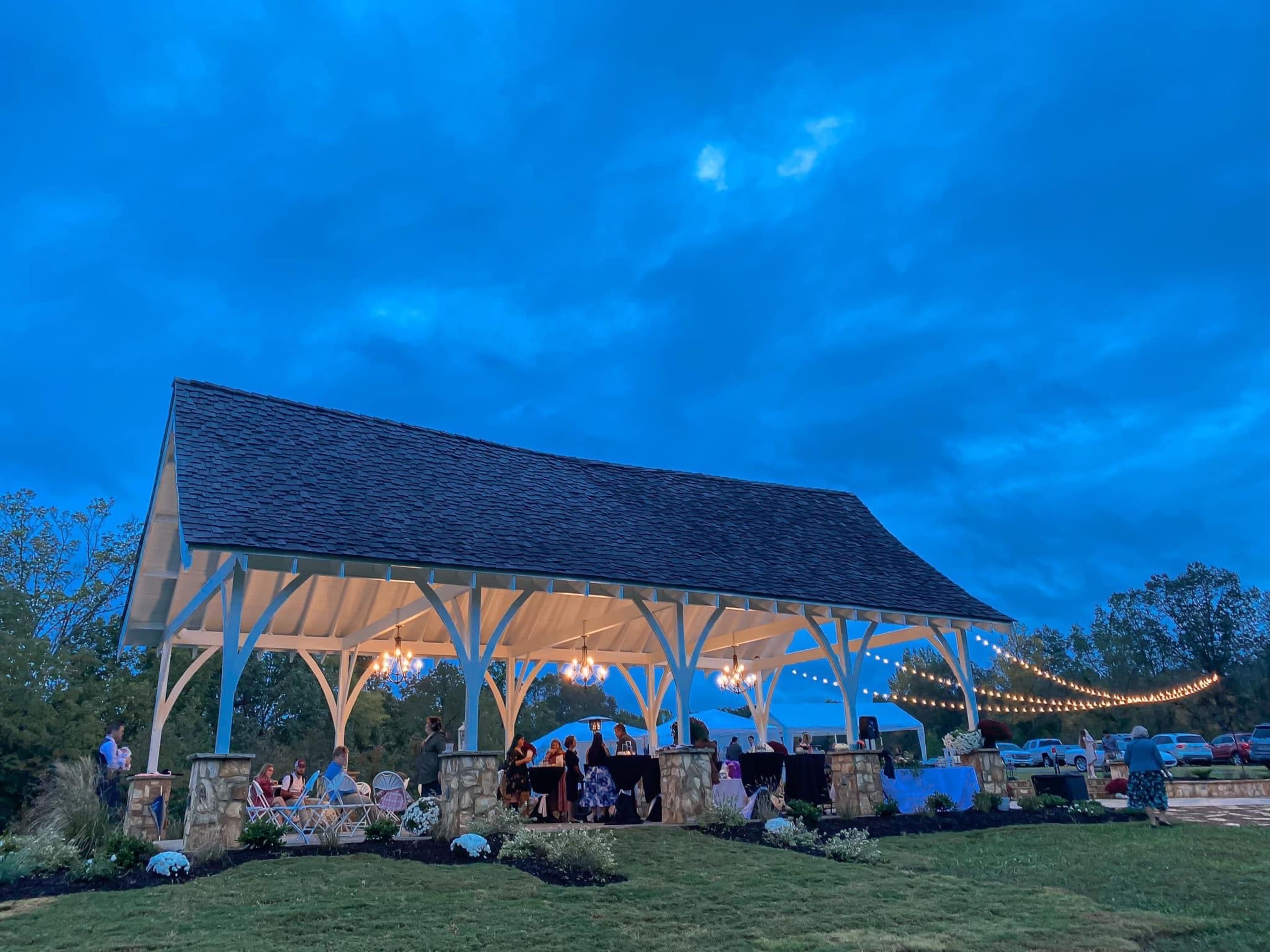 A covered pavilion with string lights and a gathering of people is set against a twilight sky.