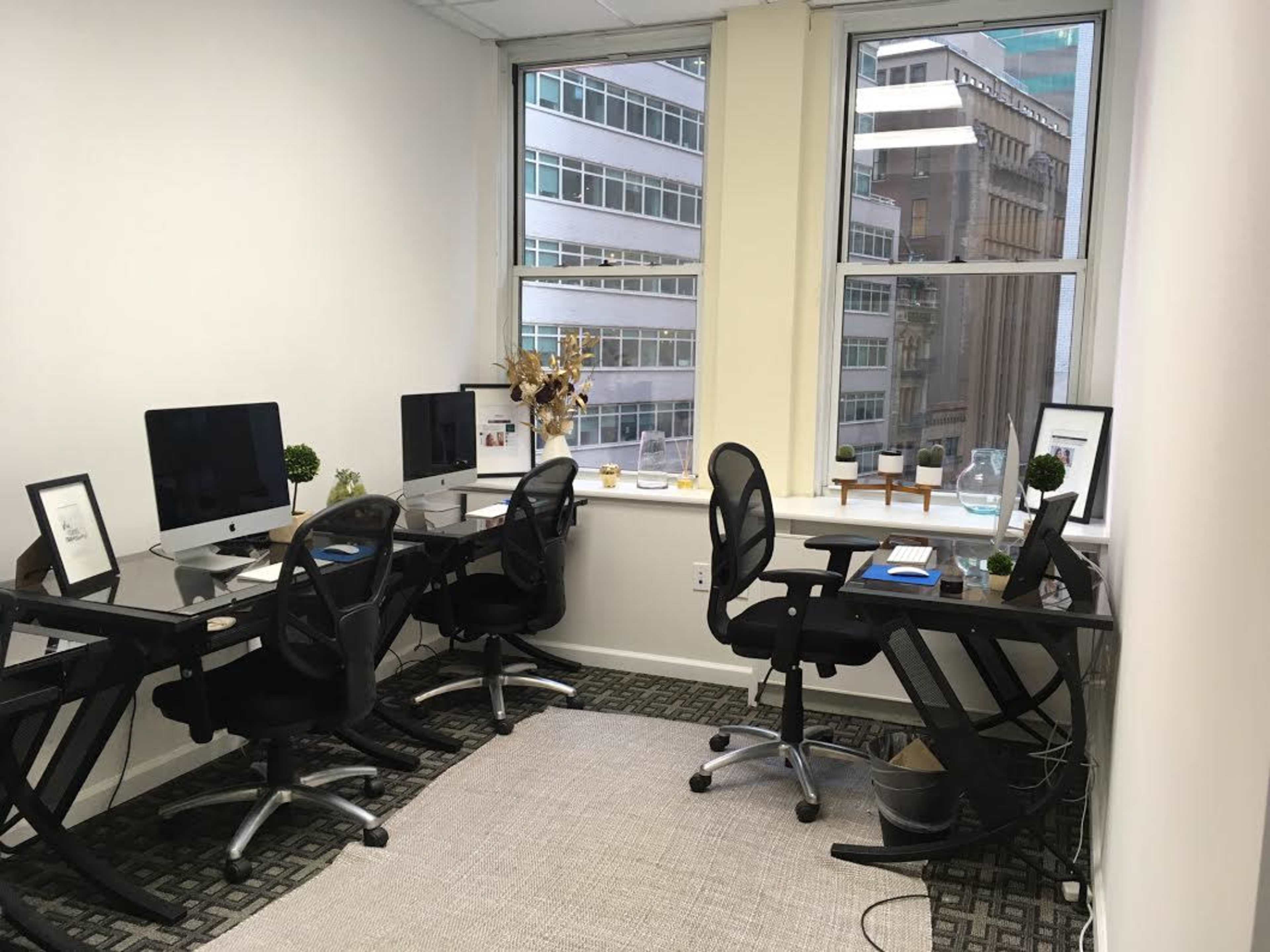 A bright office space with four workstations equipped with computers and ergonomic chairs, alongside large windows overlooking a cityscape.