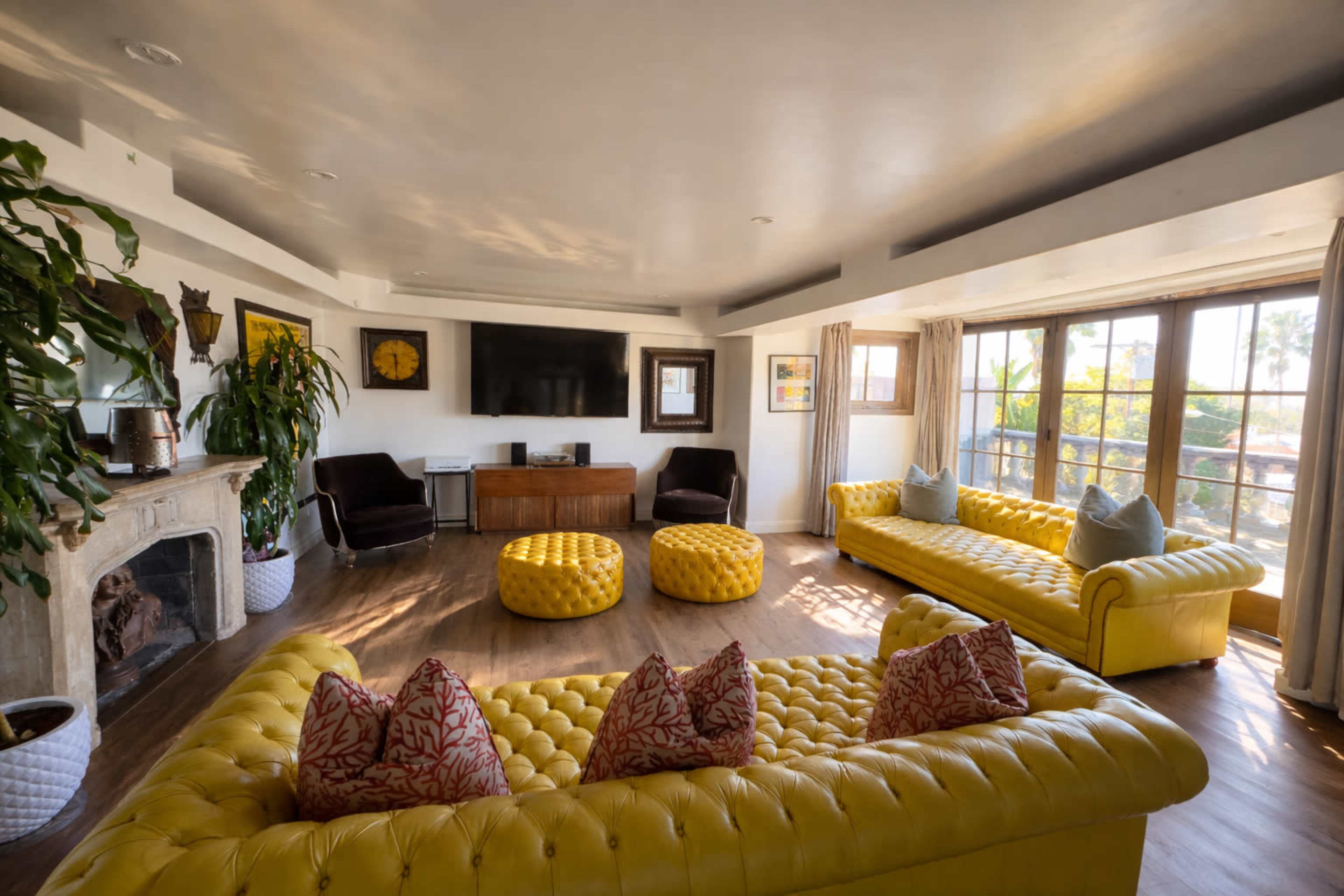 The spacious living room features yellow chesterfield sofas, round poufs, a large television, and large windows with natural light.