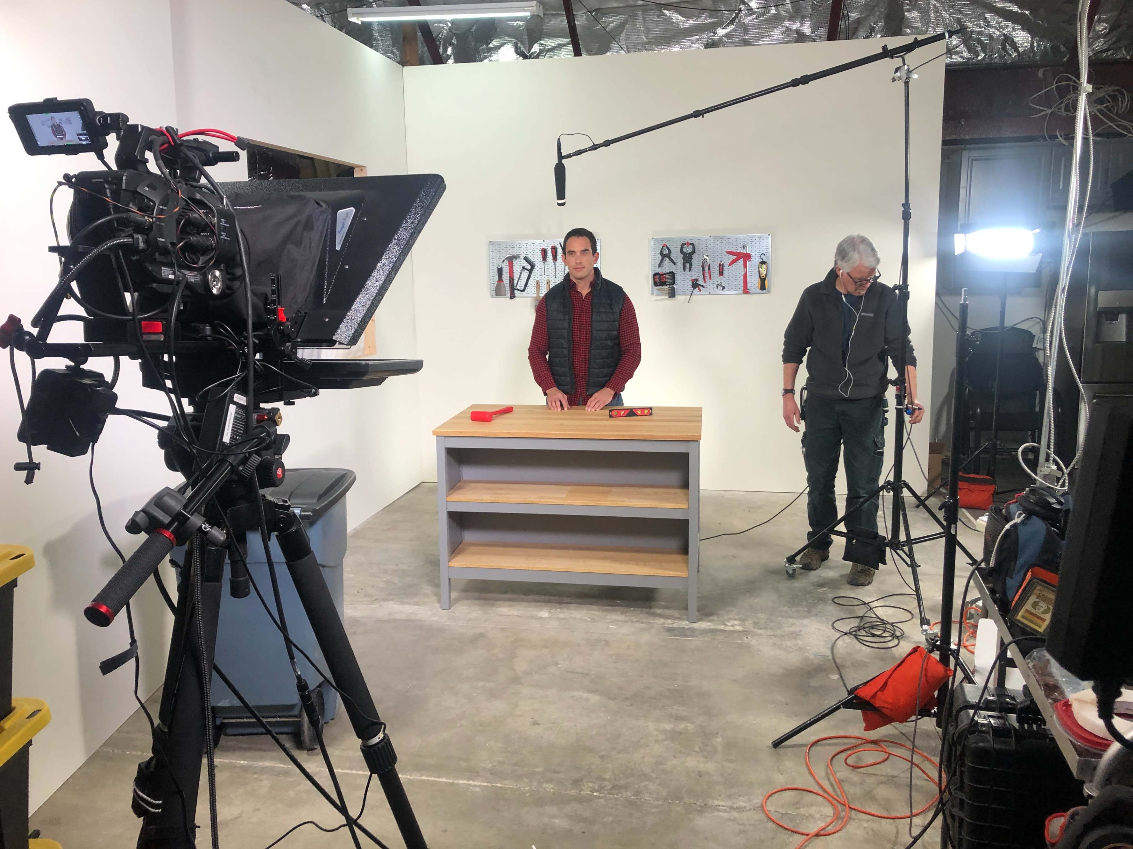 A video production setup features a man standing behind a table with tools displayed on a wall, while camera equipment and lighting are arranged around the scene.
