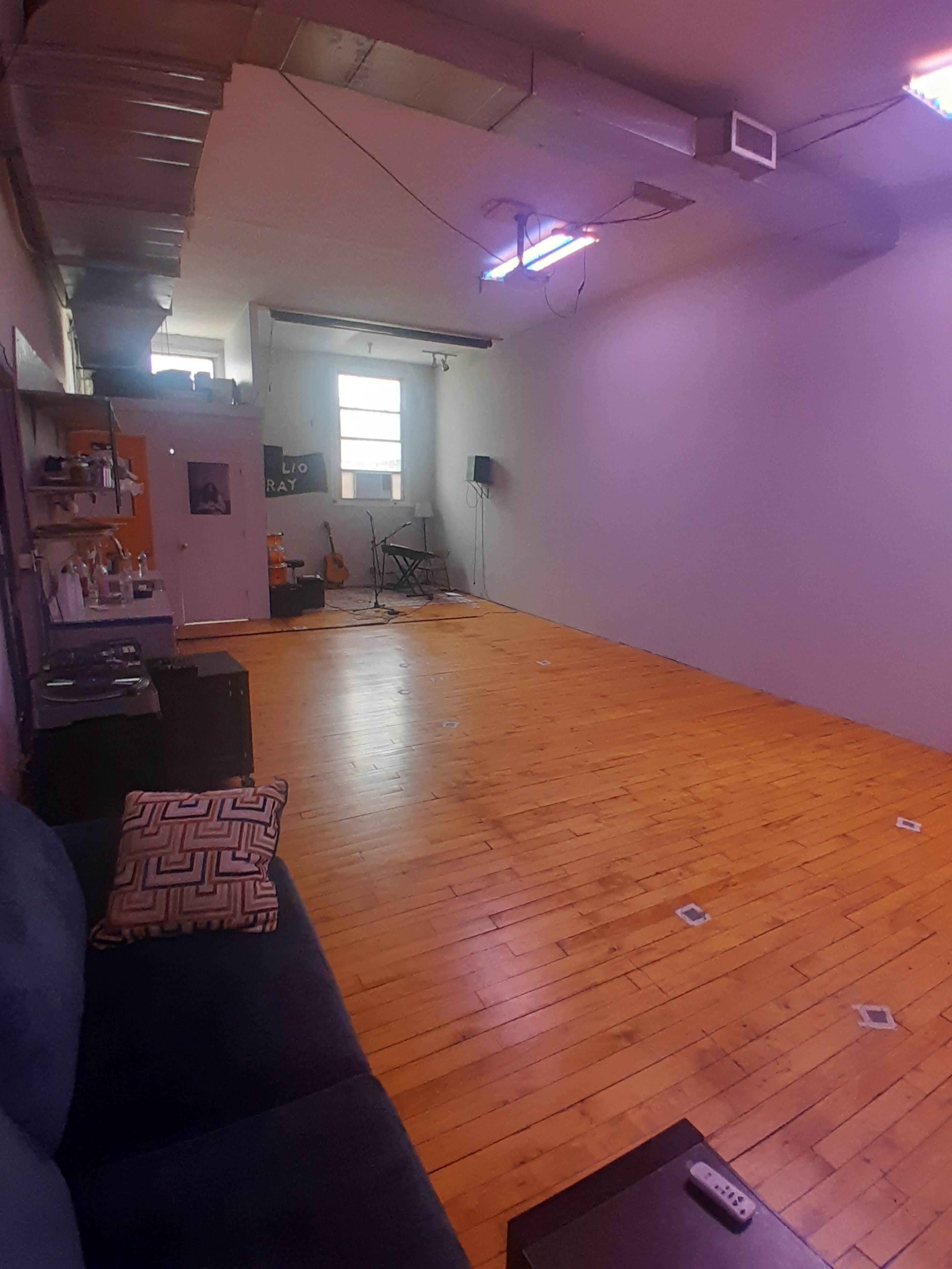 The image shows a spacious room with wooden flooring, a couch on the left, and a wall-mounted television near a window at the far end.