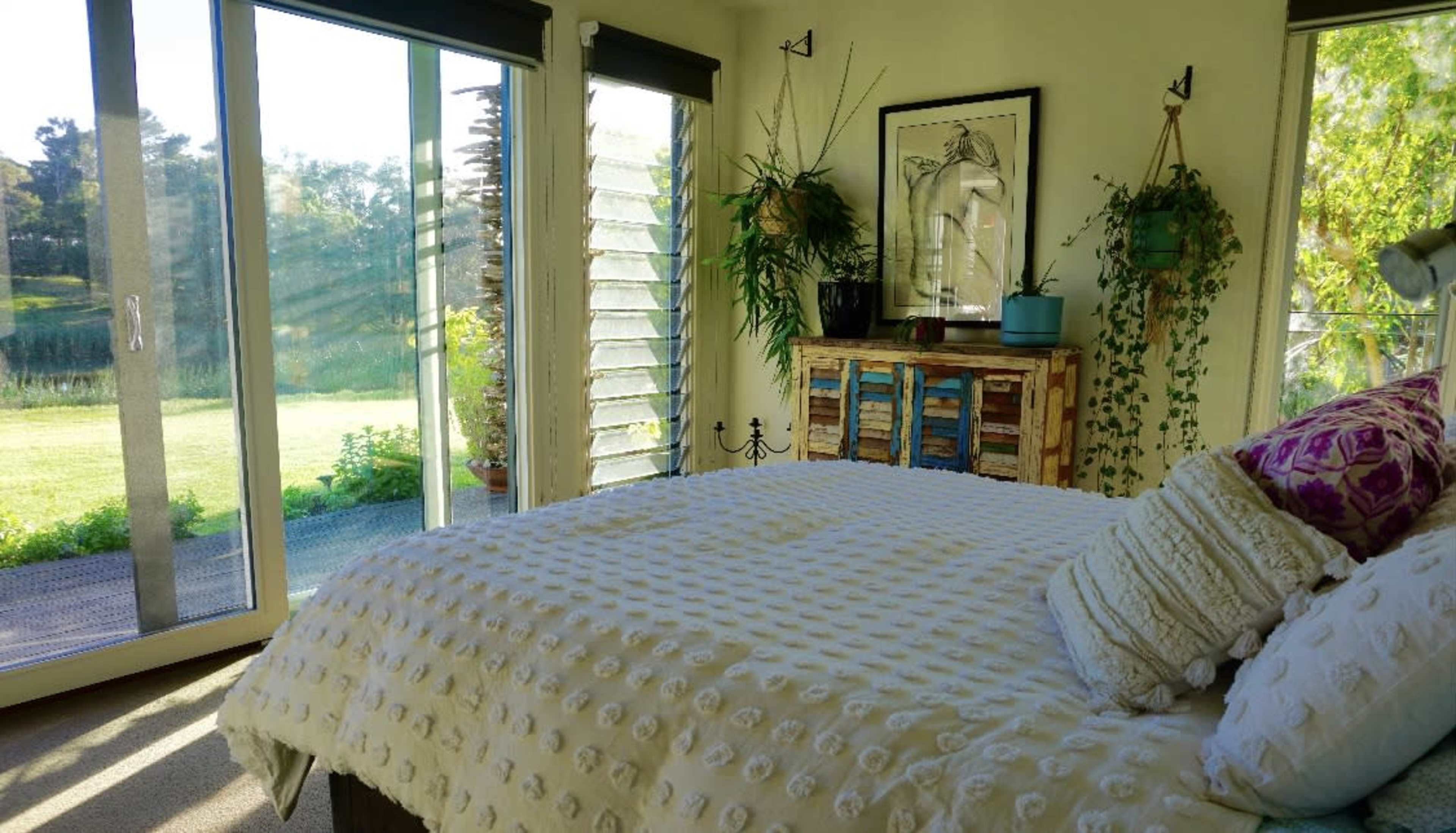 A brightly lit bedroom with a bed, large windows opening to a view of greenery, and a colorful wooden cabinet against the wall.