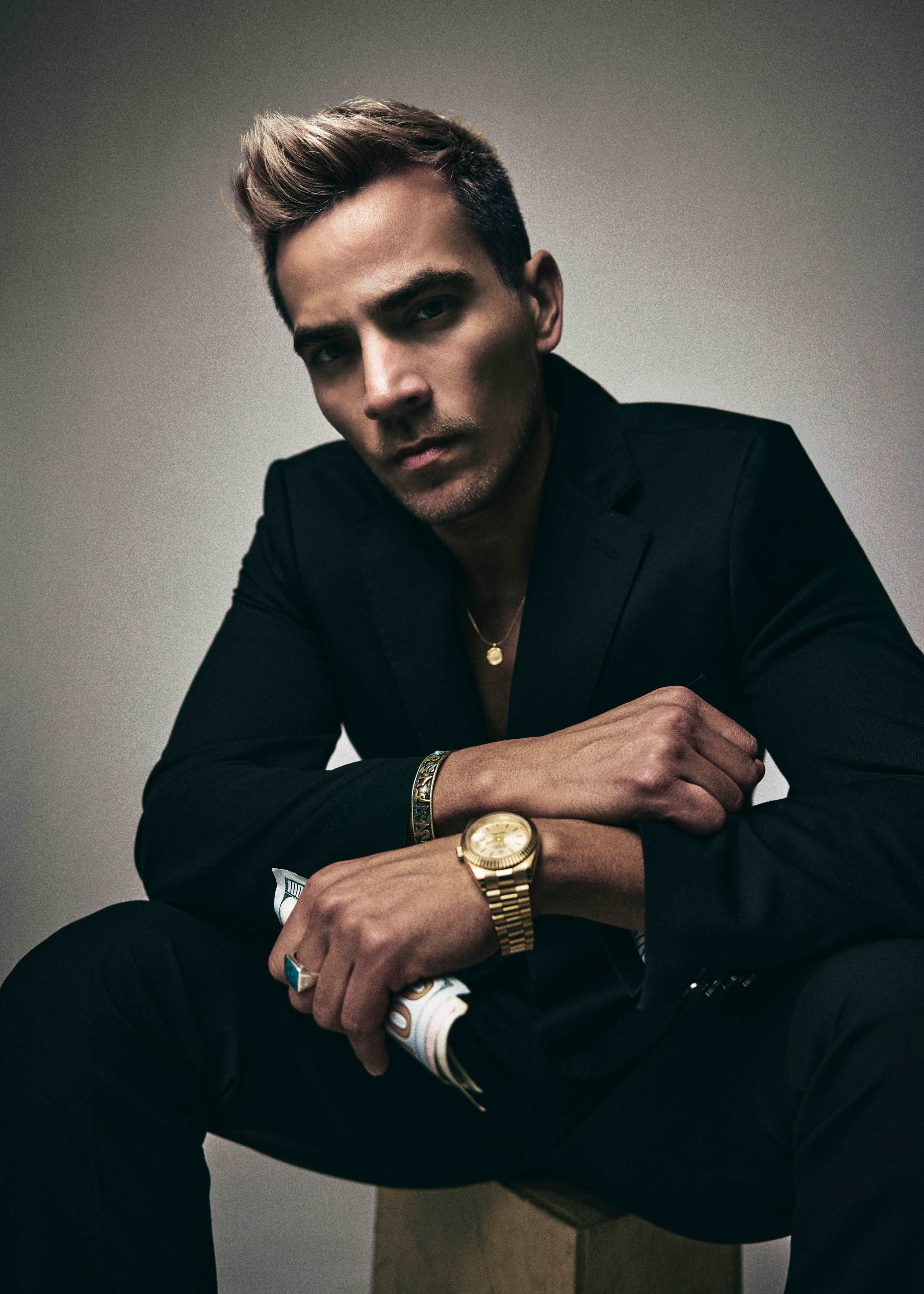 The image features a man in a black suit seated on a wooden block, looking directly at the camera with a serious expression while wearing a gold watch and holding a magazine.