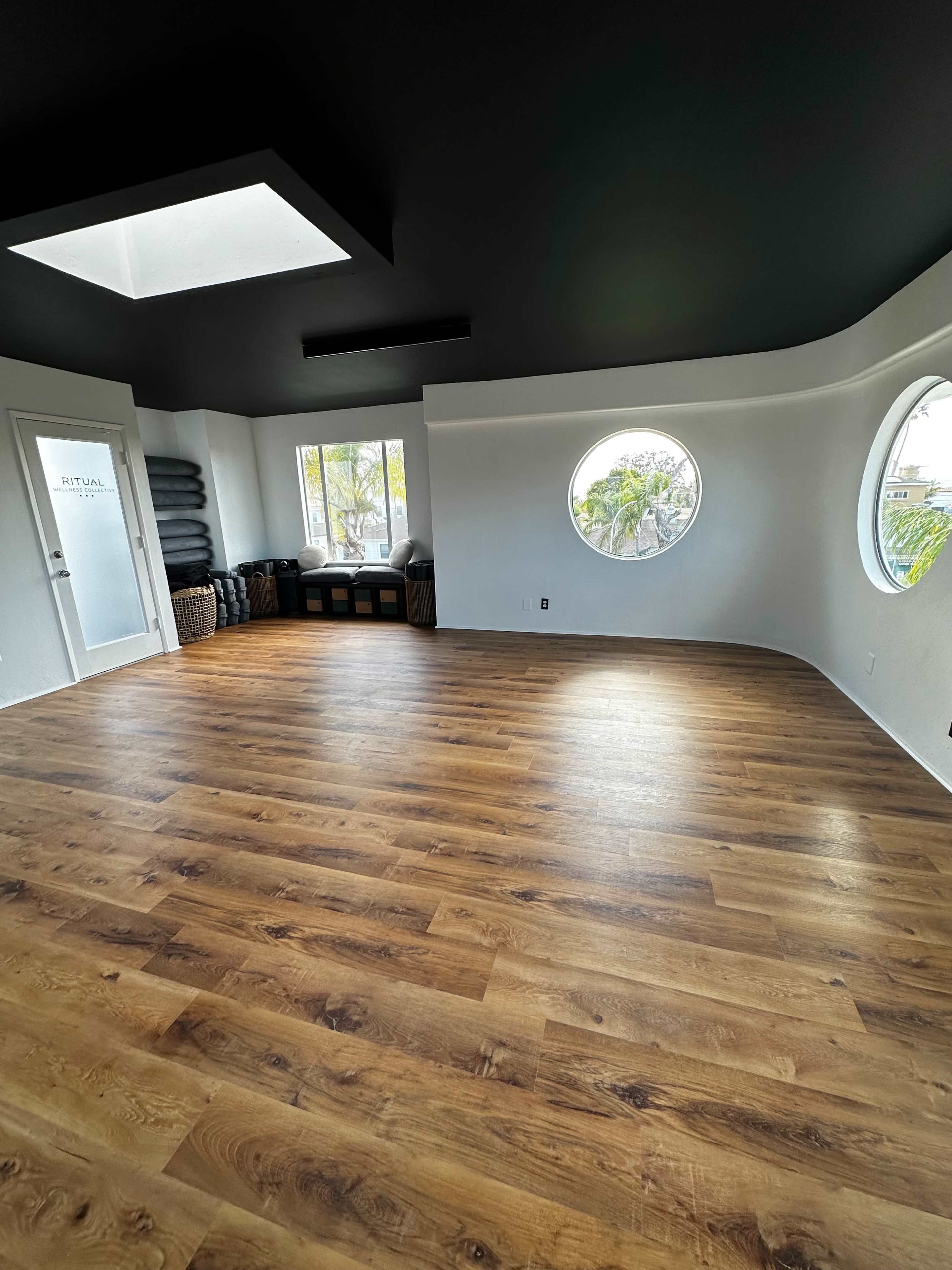 The image shows a spacious room with wooden flooring, a black ceiling, and circular windows providing natural light.