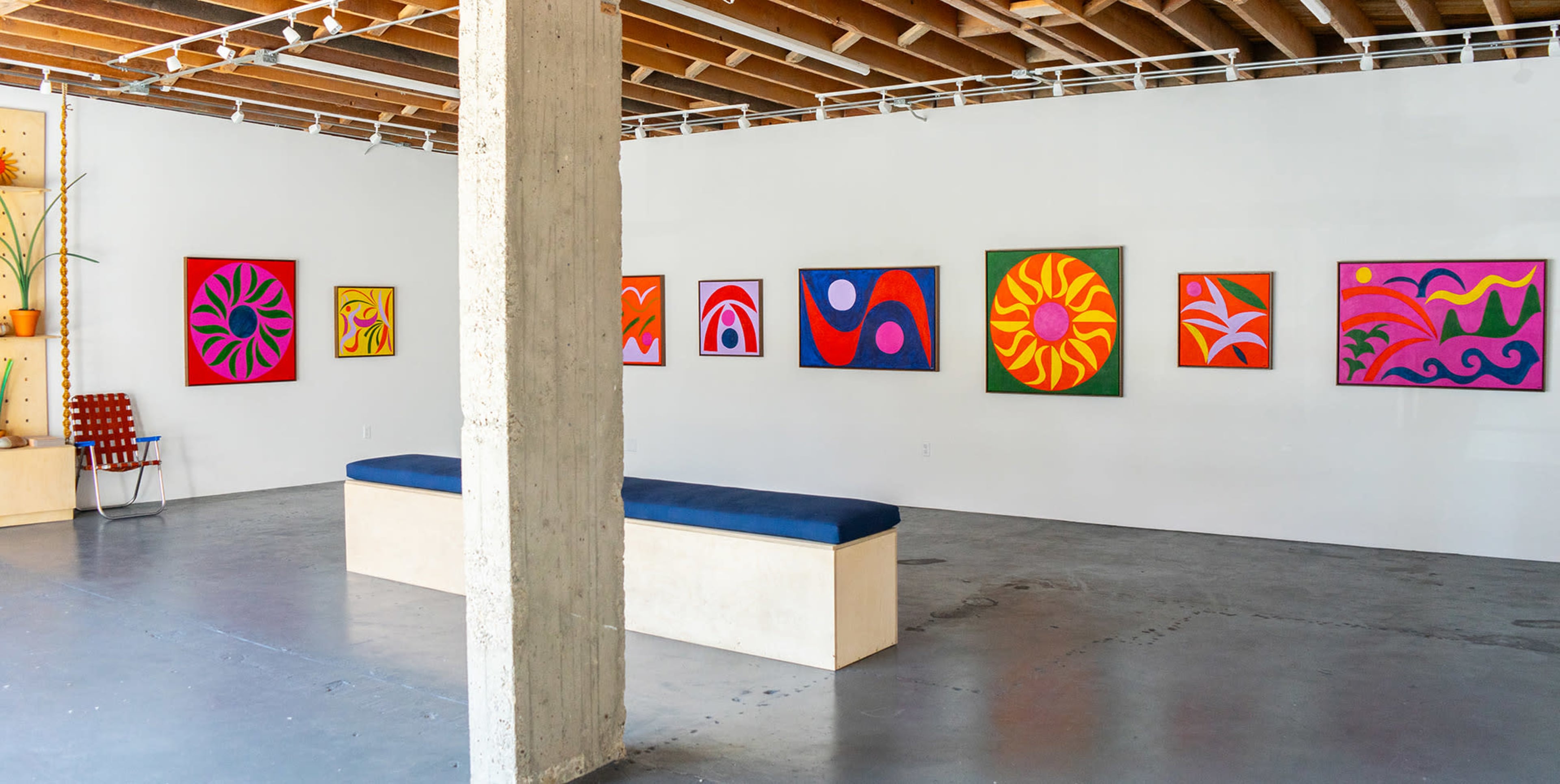 The image shows an art gallery with colorful abstract paintings displayed on the white walls and a long bench in the center of the room.