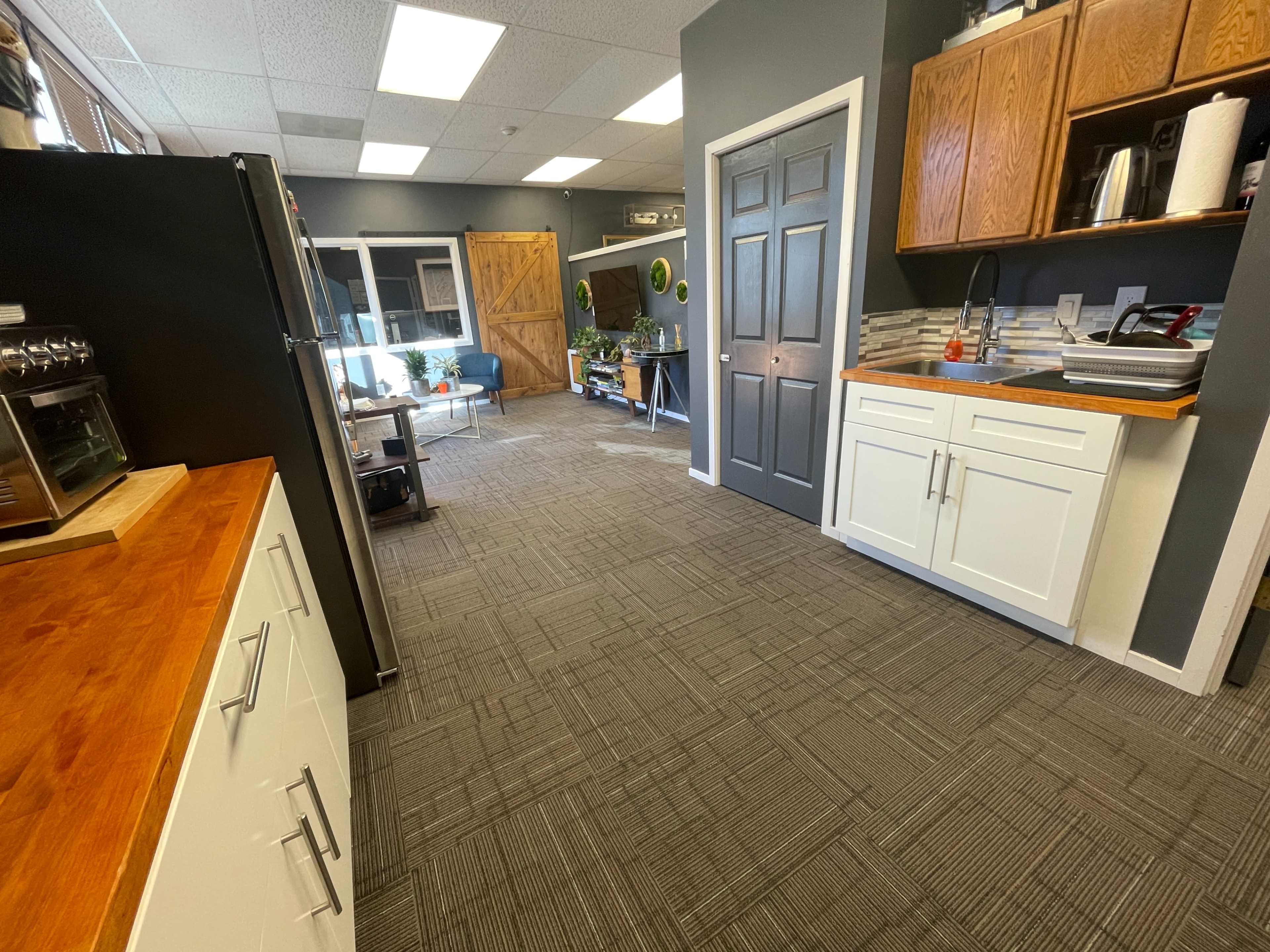 The image shows a spacious interior of an office or workspace with a kitchen area on the left, hardwood cabinetry, and an open seating area in the background.