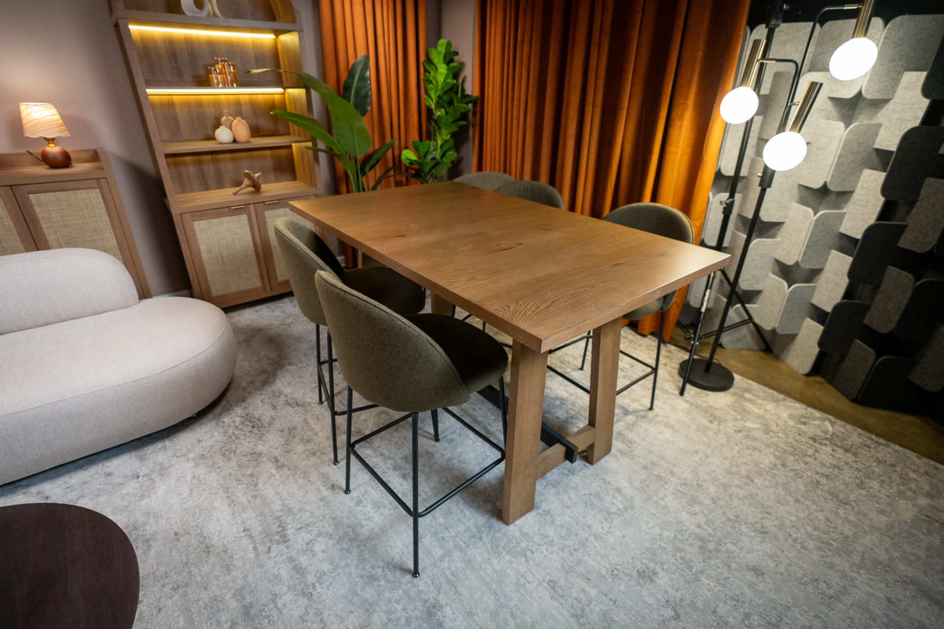 A modern dining area features a rectangular wooden table surrounded by upholstered chairs under warm lighting, with a backdrop of orange curtains and decorative plants.