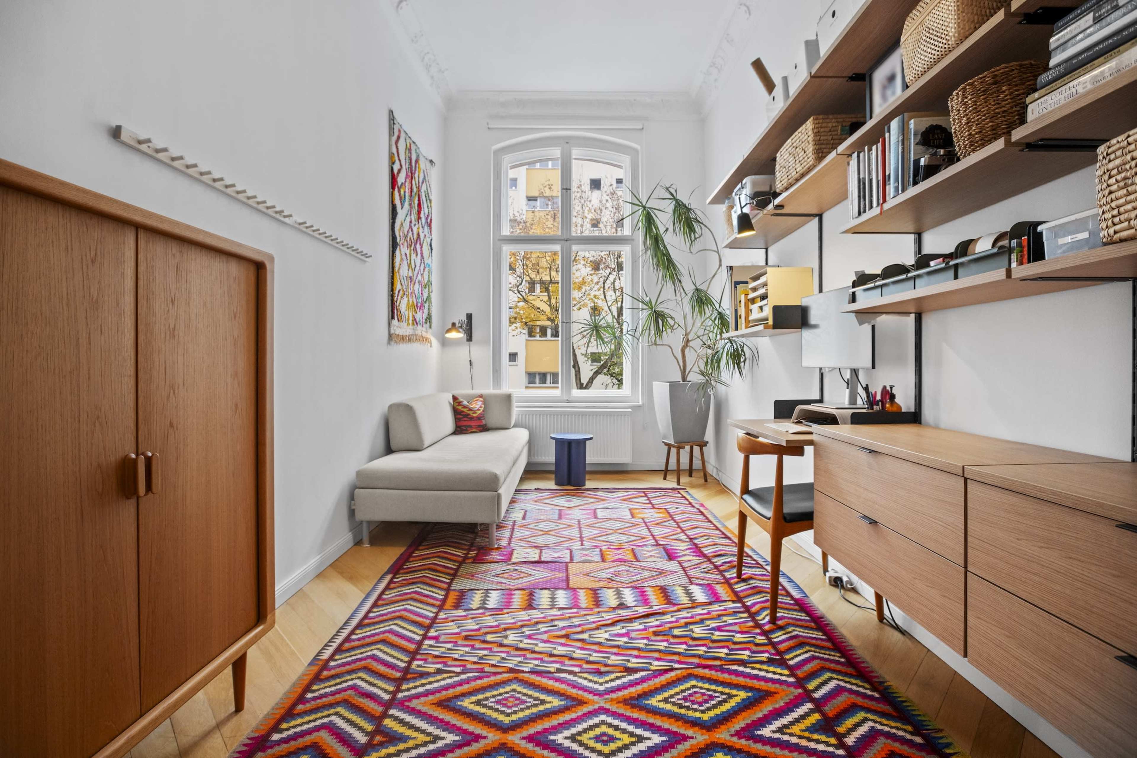 The image shows a modern, minimalistic workspace with a gray sofa, a wooden desk, shelves, and a colorful patterned rug.