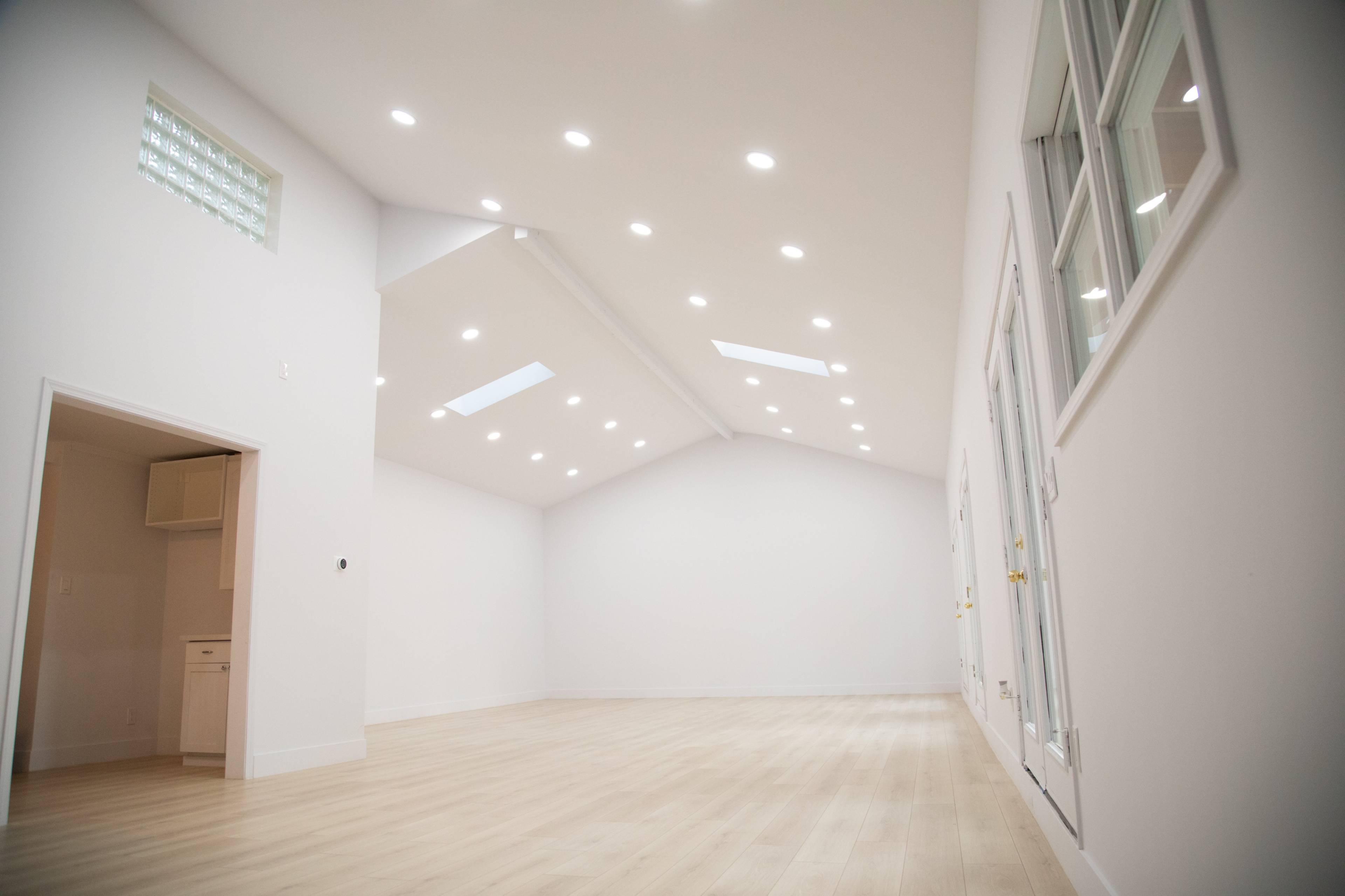 A spacious interior room features a high ceiling with recessed lighting and white walls, complemented by large windows and a light wooden floor.