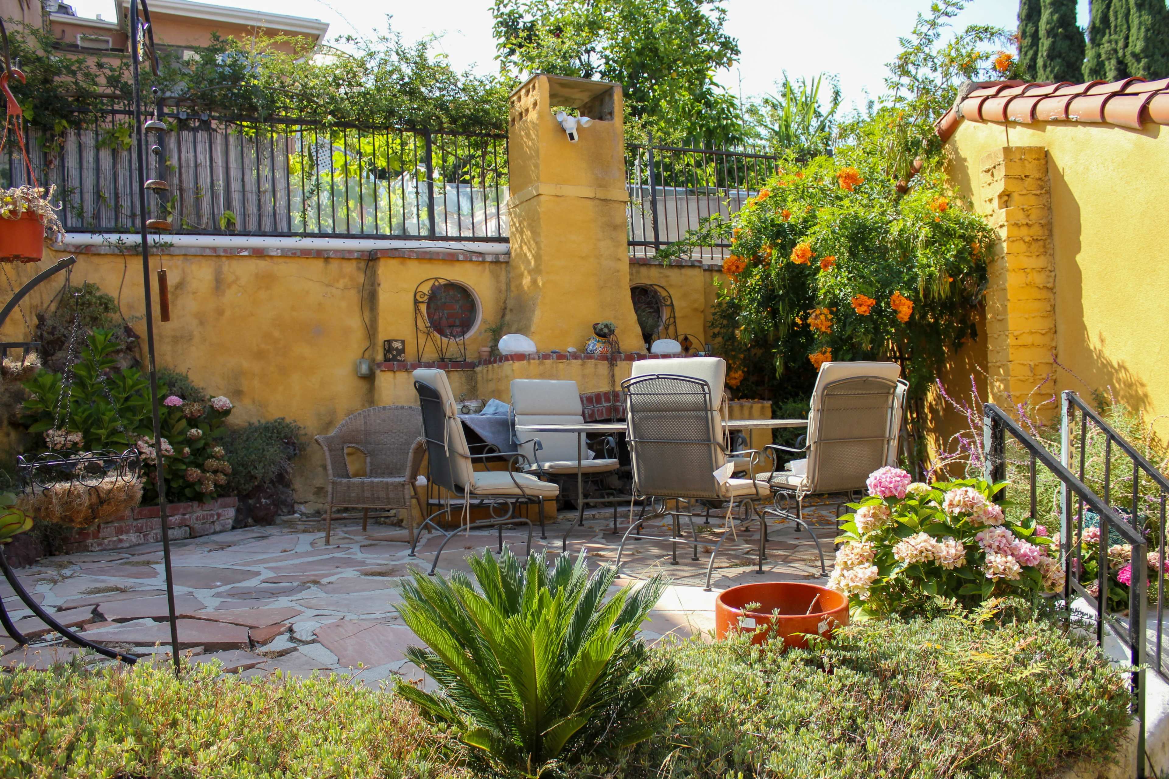 The image shows a stone patio in a garden surrounded by colorful flowers and plants, with a table and chairs arranged for seating.