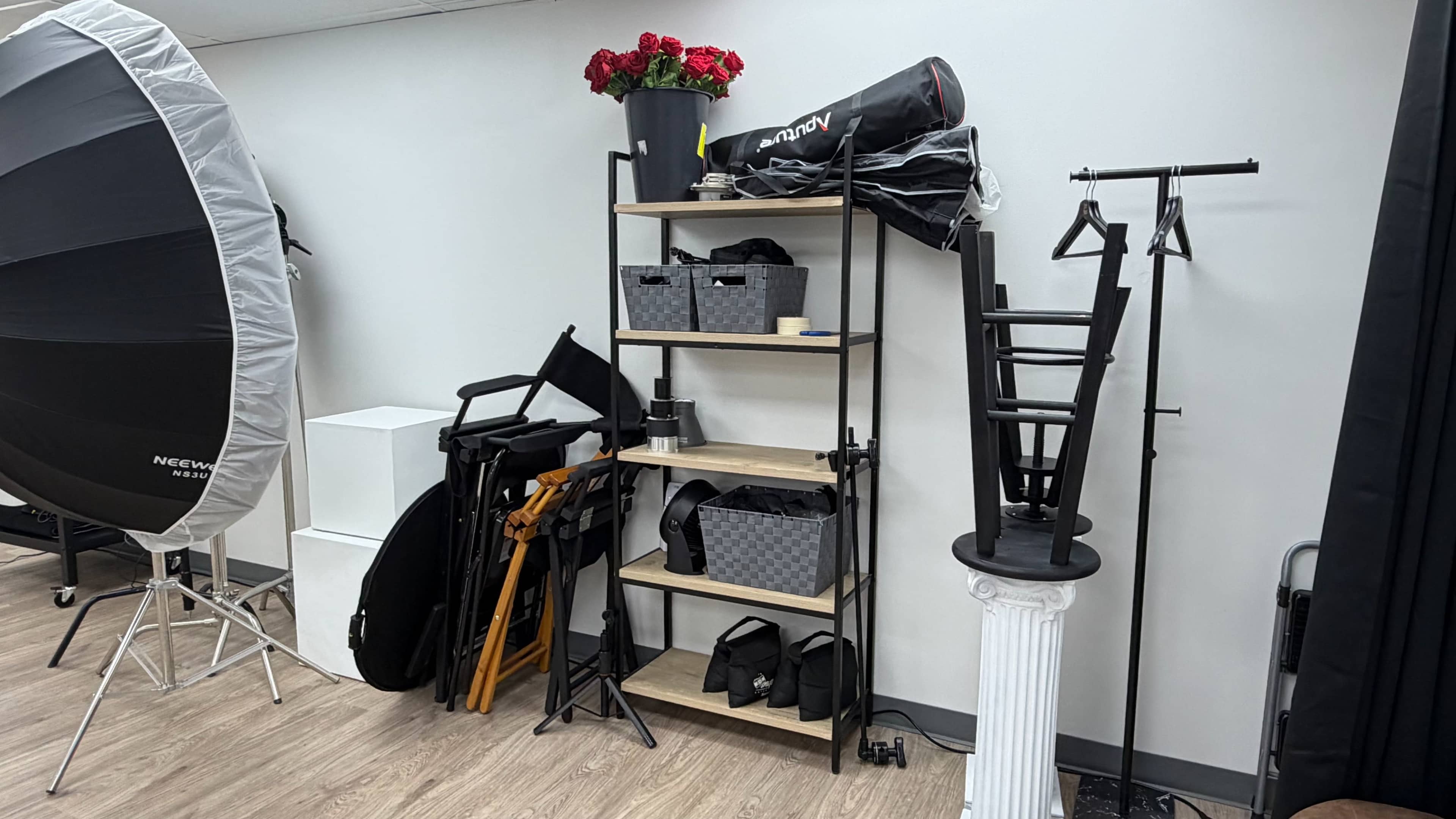 The image shows a studio space with a shelving unit displaying photography equipment, including bags, a flower pot, and various stands alongside a large lighting umbrella.