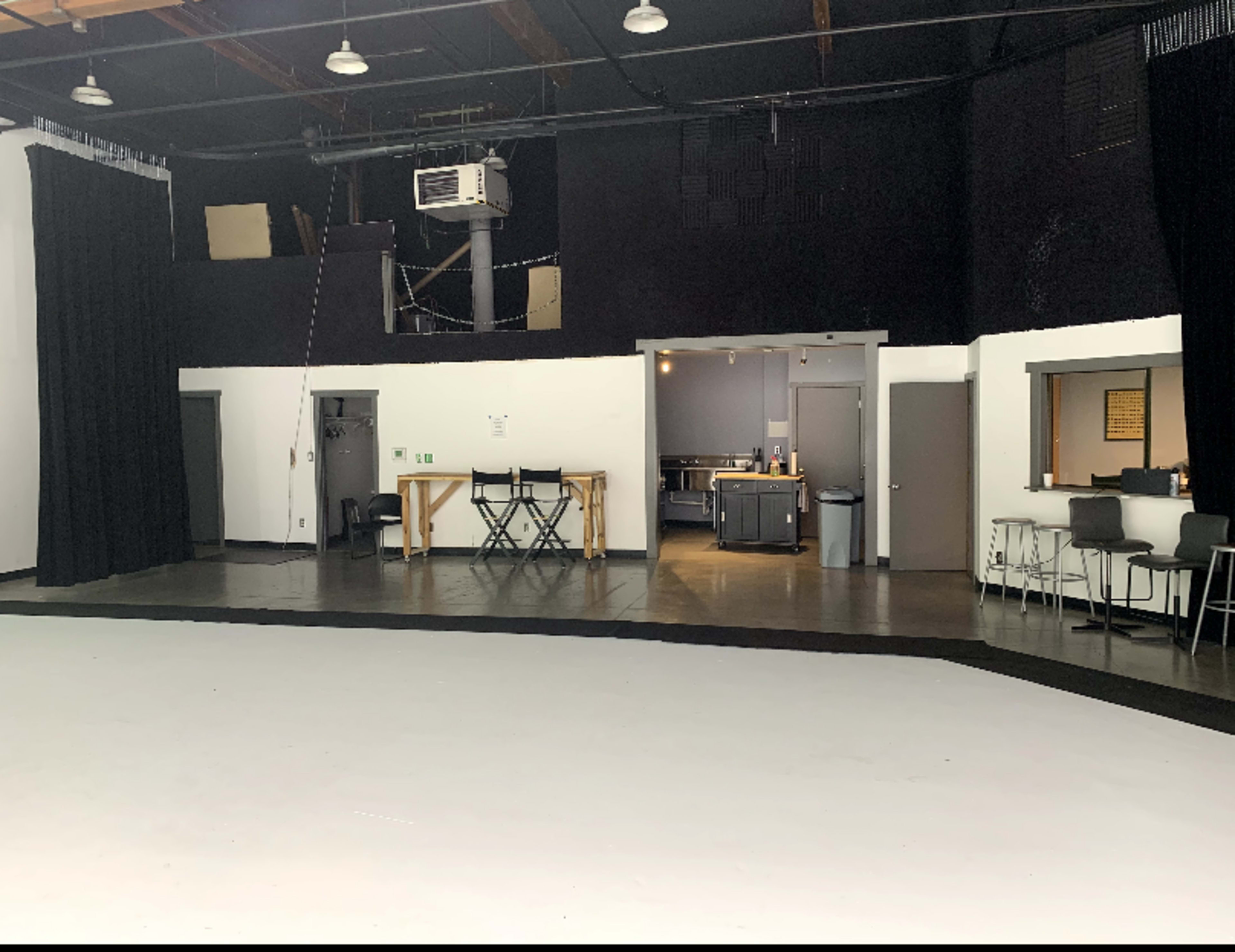 The image shows an empty studio space with black walls, a white floor, and an area with kitchen amenities and seating.