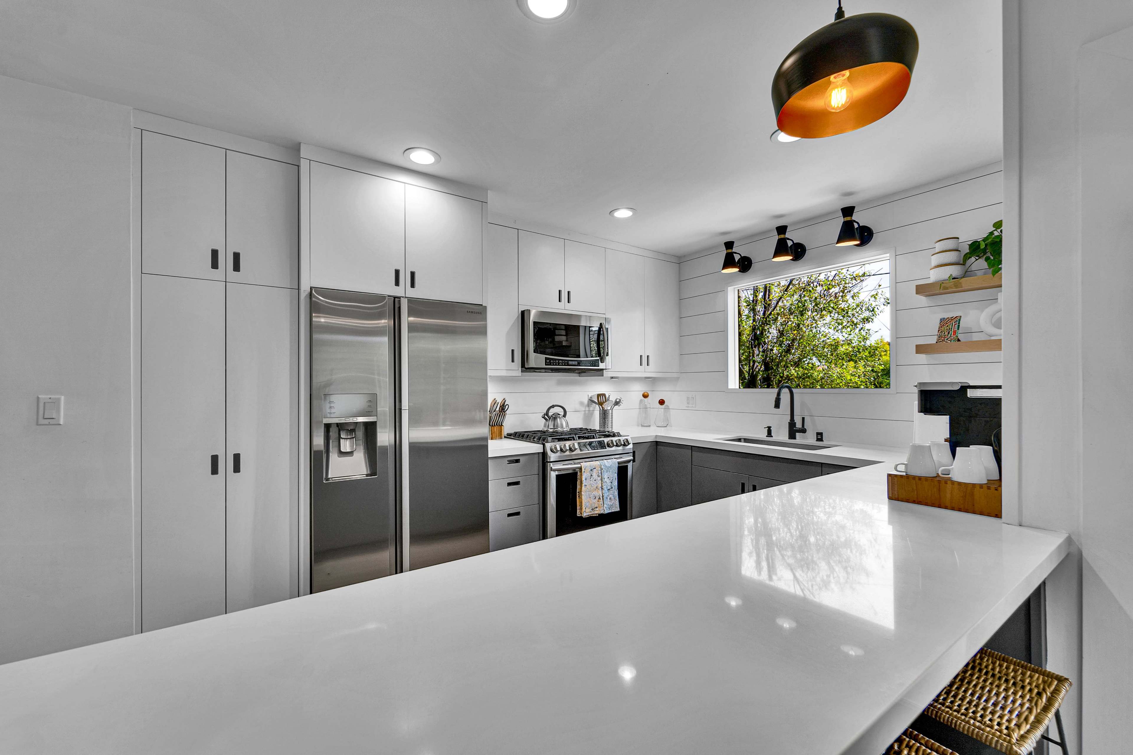 The image shows a modern kitchen with white cabinets, stainless steel appliances, and a countertop overlooking a window with greenery outside.
