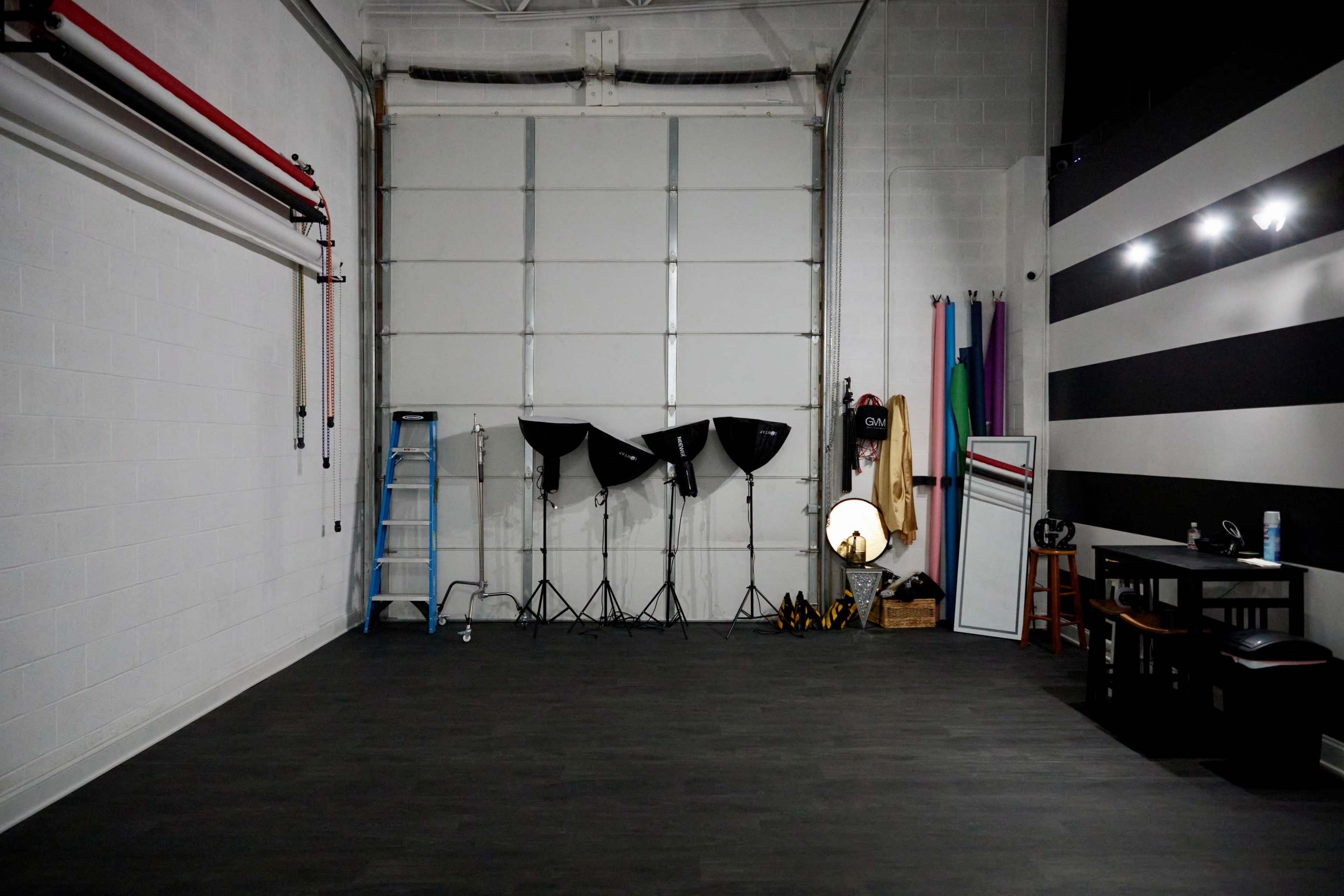 The image shows a spacious studio with a large garage door, lighting equipment, a ladder, and various props set against a wall with black and white stripes.