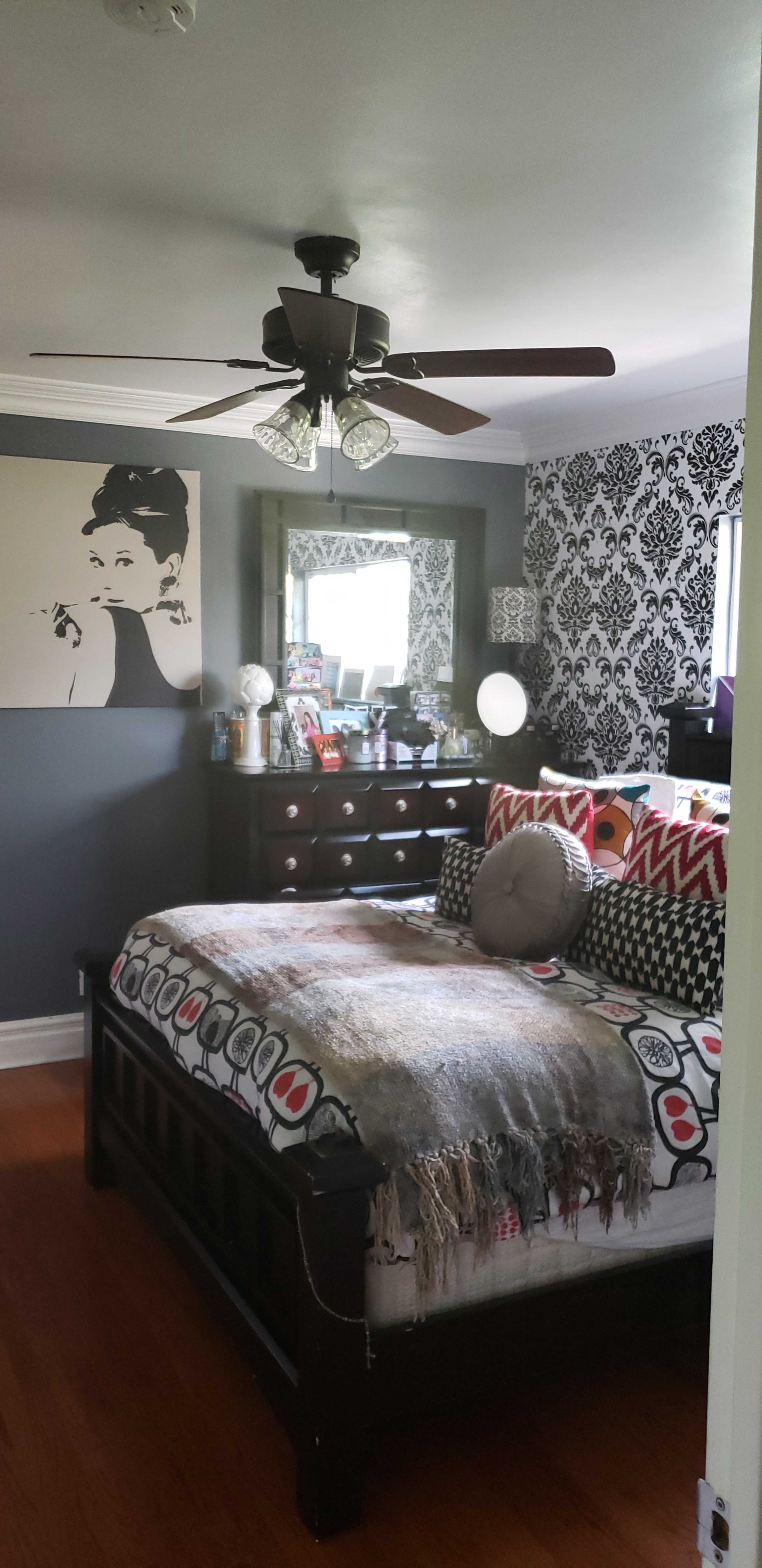 A bedroom features a dark accent wall, a large bed with a patterned blanket, a dresser with multiple drawers, and a decorative mirror, alongside a wall art piece.