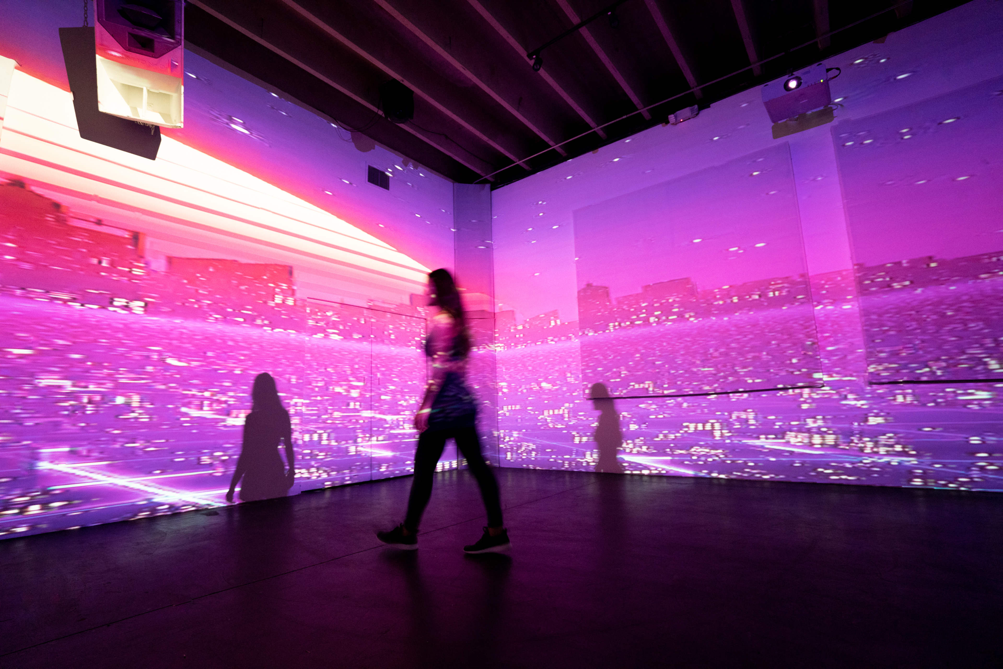 A person walks through a dimly lit room where vibrant digital projections of a cityscape are displayed on the walls.