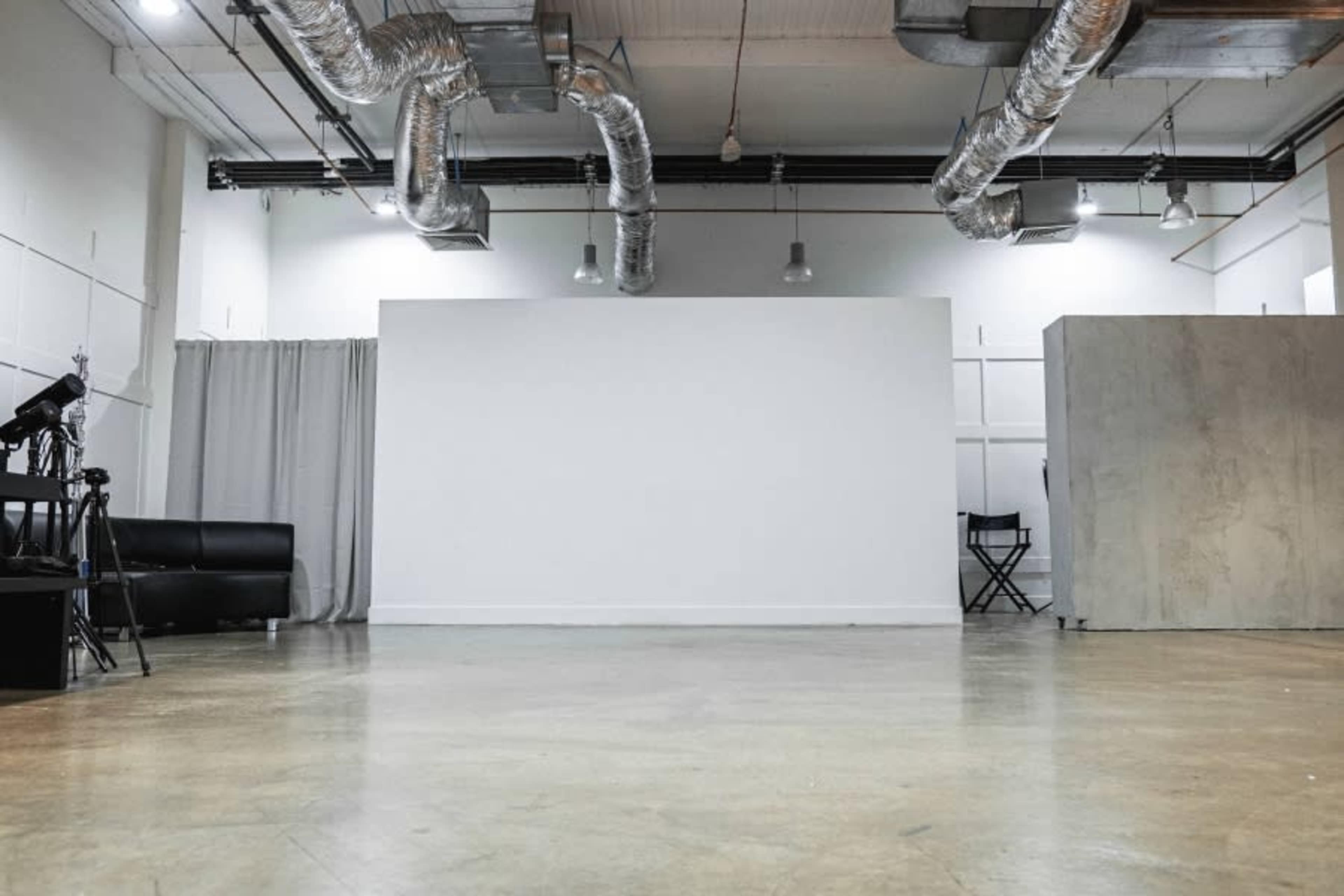 A spacious, empty studio with metal ductwork overhead, a black couch, a director's chair, and a gray partition wall.