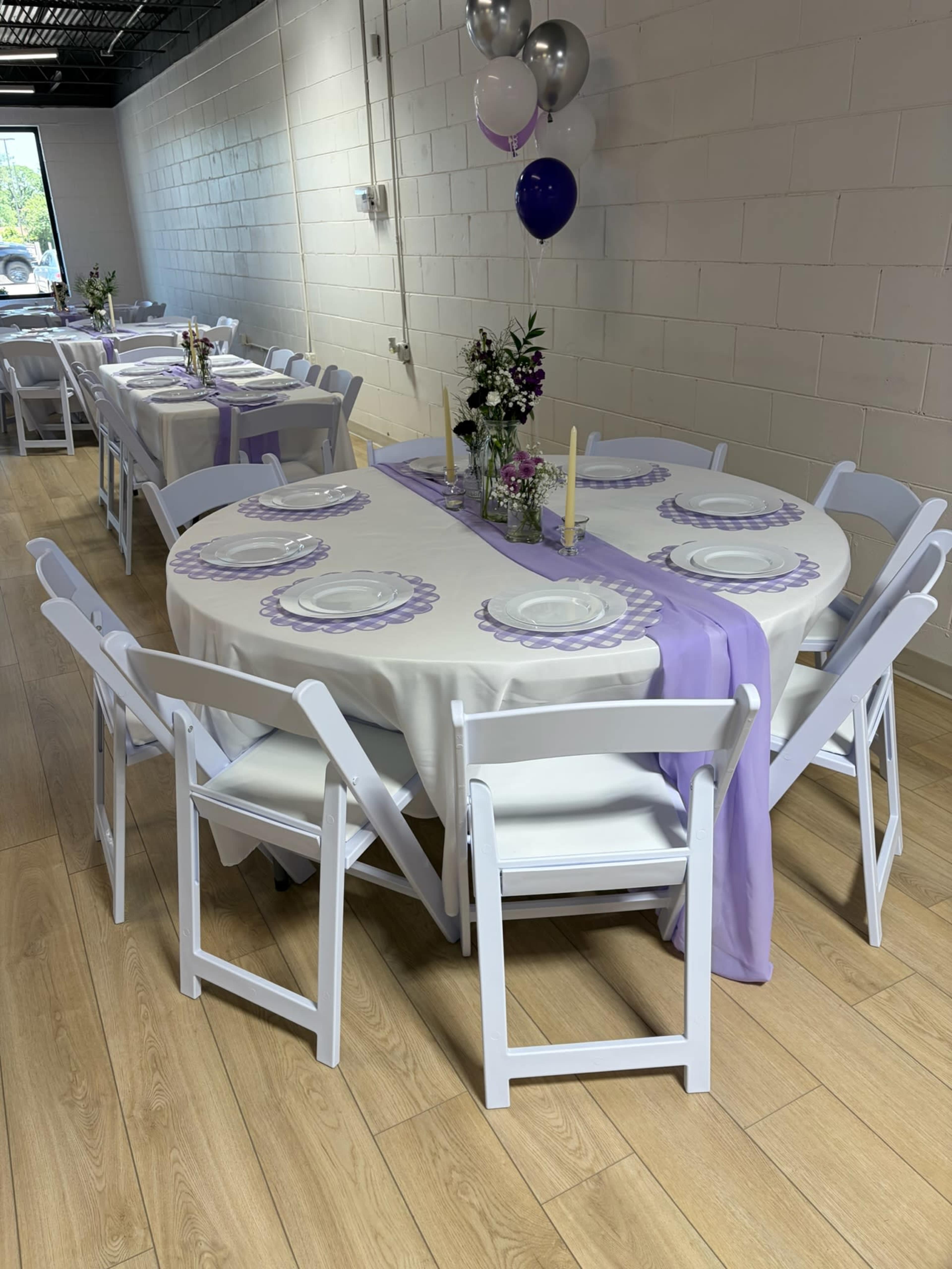 The image shows a dining area set with several tables covered in white and purple tablecloths, arranged with plates, floral centerpieces, and decorated with balloons.