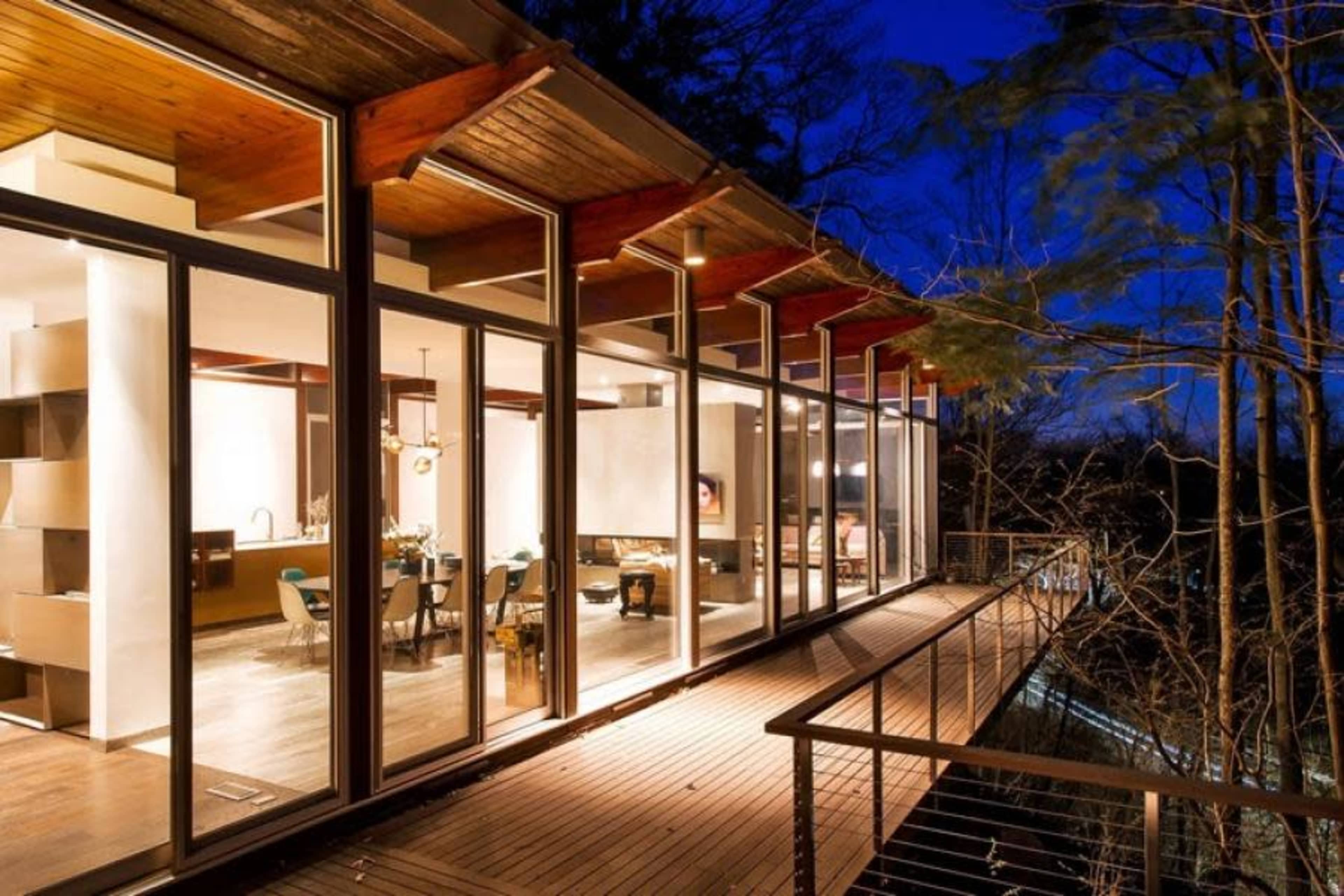 A modern wooden deck extends from a glass-walled house, surrounded by trees at dusk.