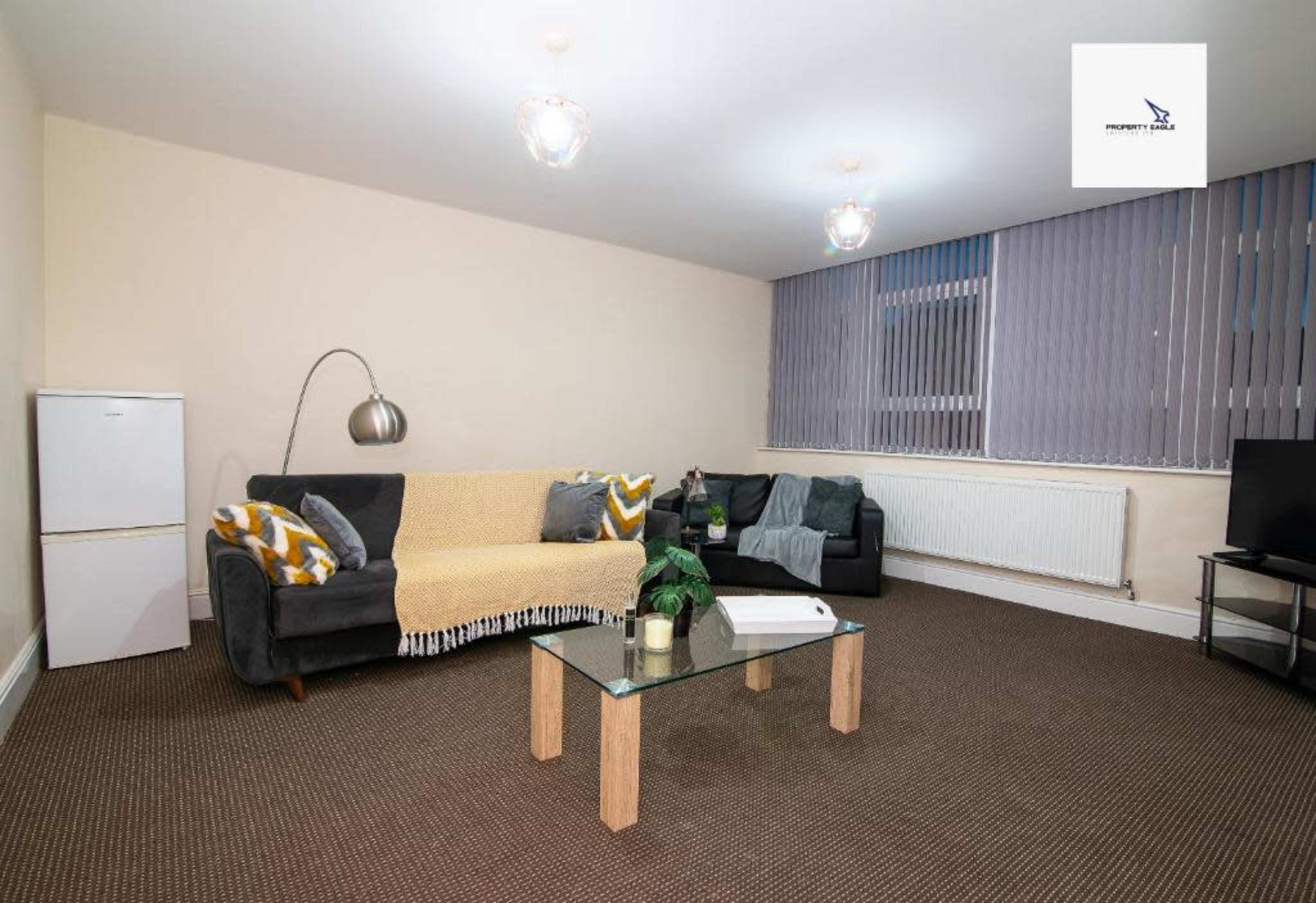A minimalist living room features a gray couch with a yellow throw, a glass coffee table, a white refrigerator, and large windows with vertical blinds.