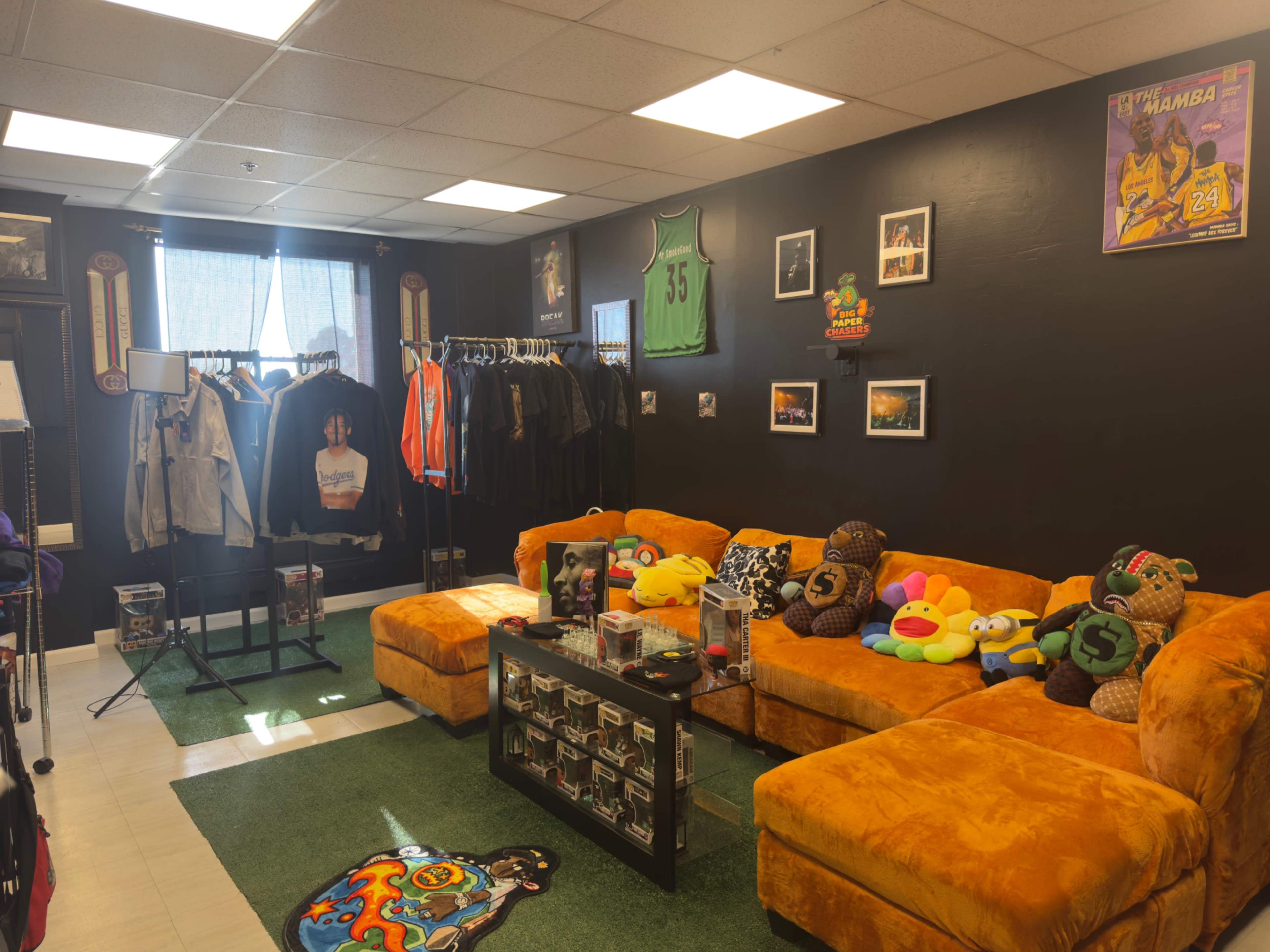 The image shows a cozy room featuring orange couches, a clothing rack with various garments, and collectibles on a glass coffee table amidst a green carpet.