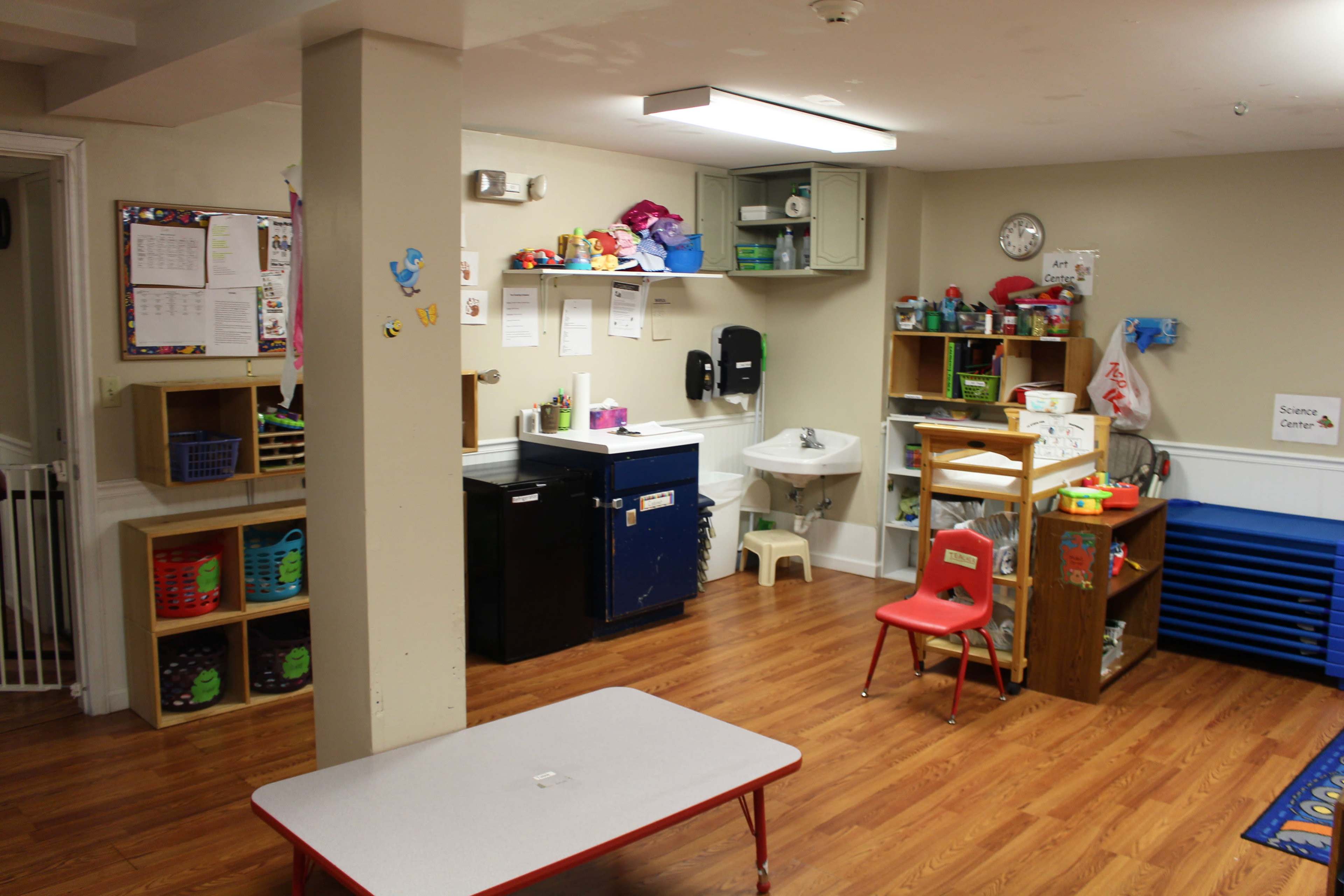 A brightly lit basement room set up as a play area, featuring storage shelves, a small table, a kitchenette with a sink, and various toys and materials organized within the space.