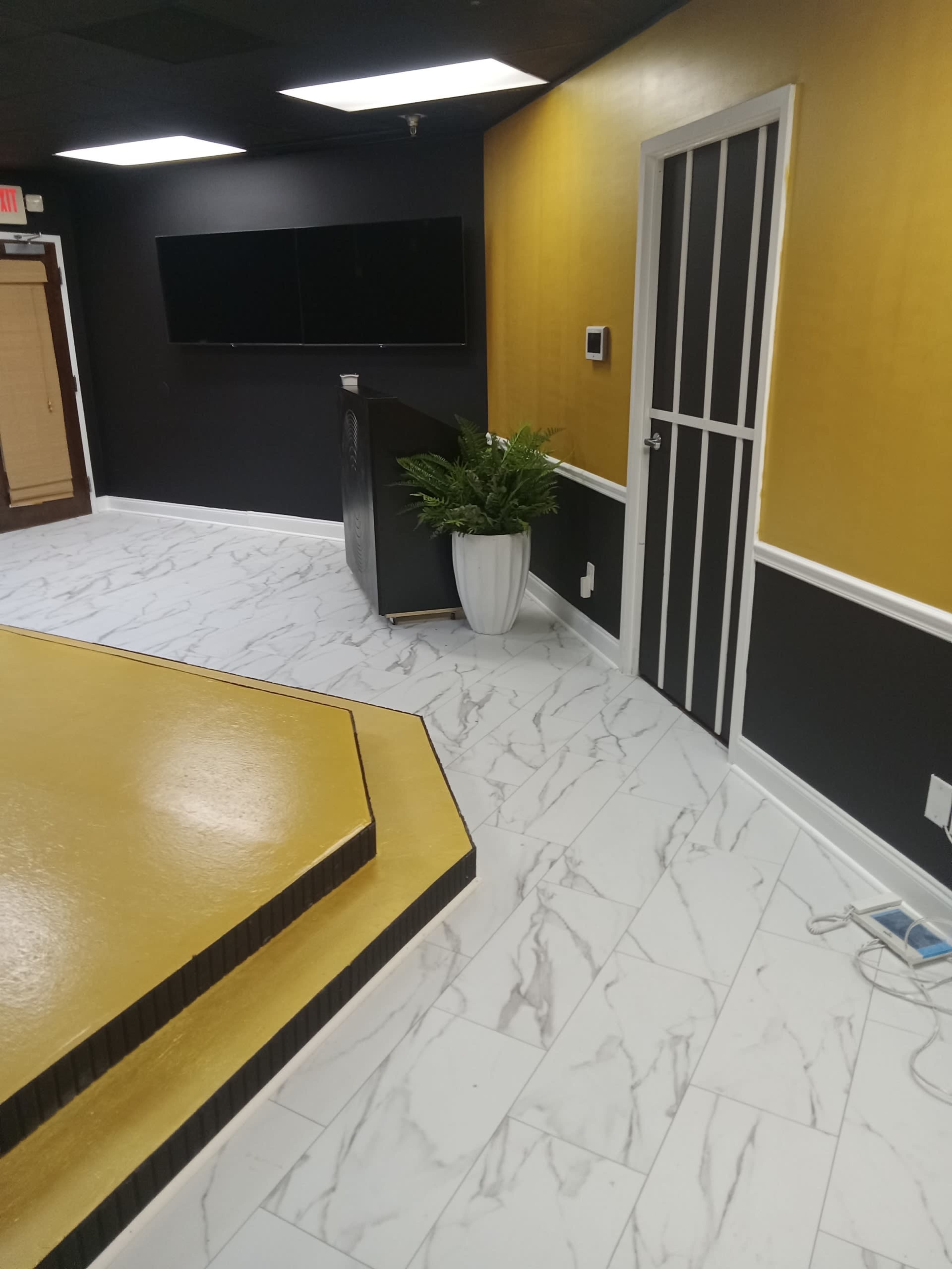 The image shows a modern room with black and yellow walls, a podium with a plant, and a wall-mounted screen in the background.