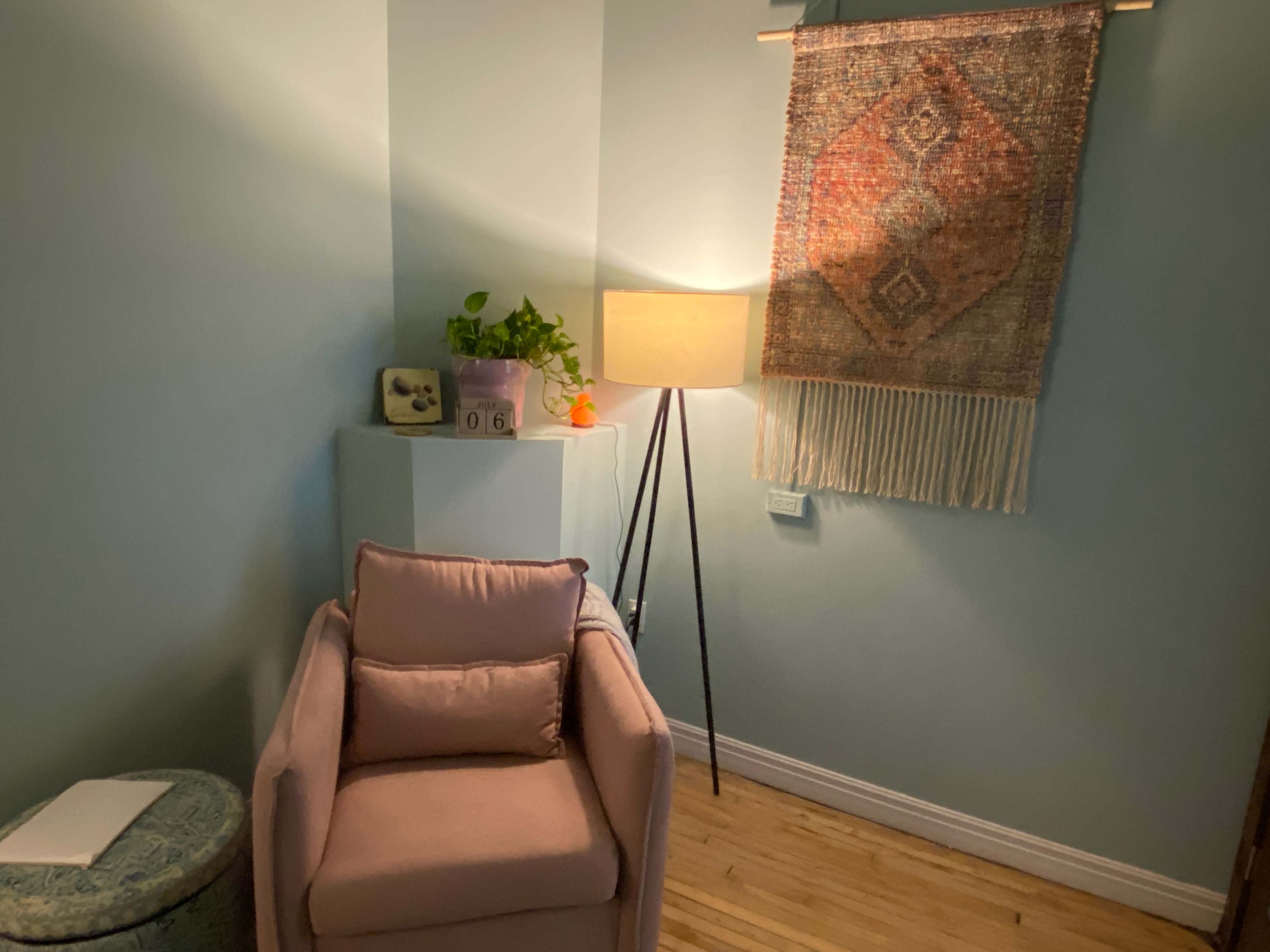 A cozy corner of a room featuring a pink armchair, a lamp on a tripod stand, a small plant, and a decorative wall hanging.