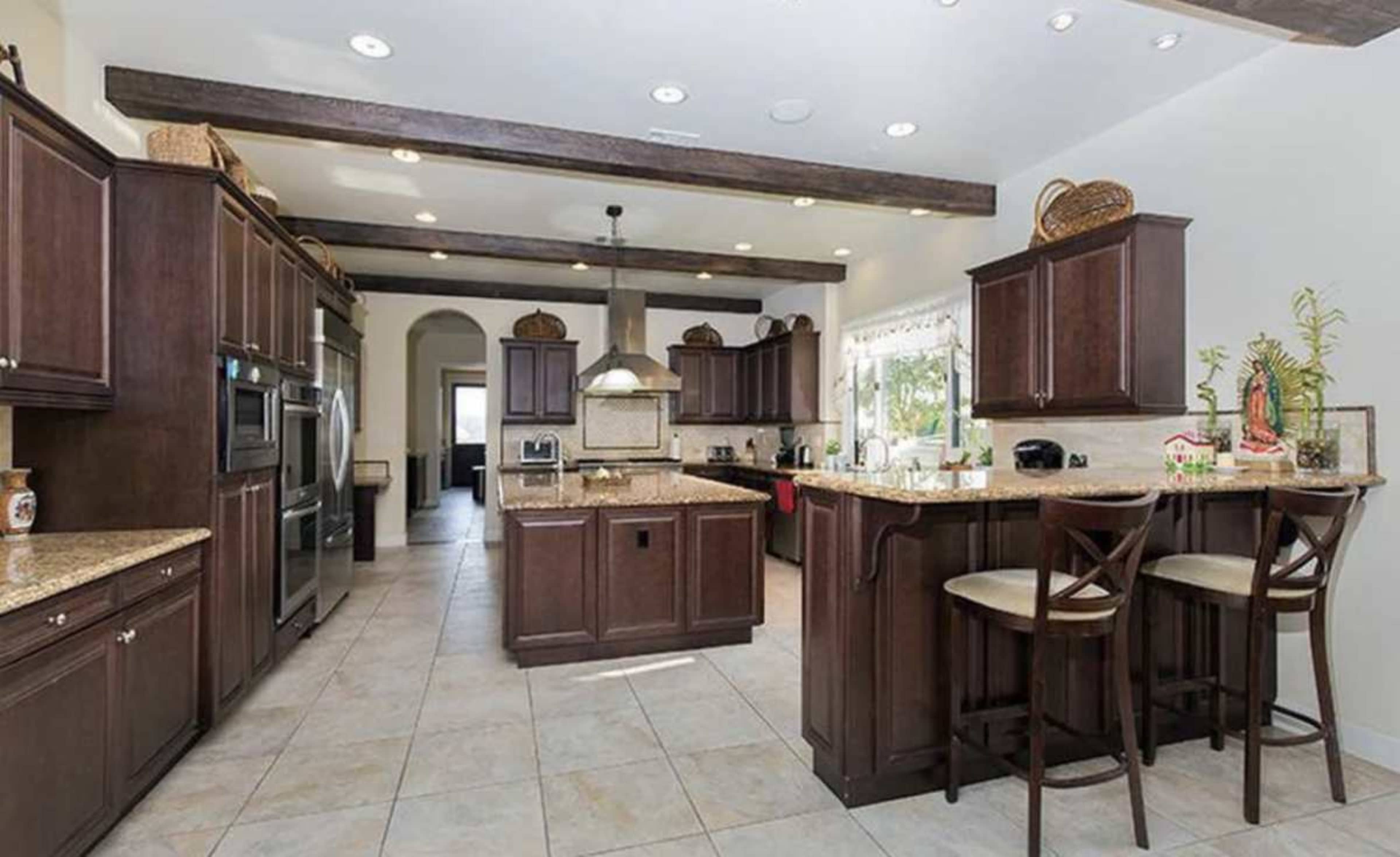 The image shows a spacious kitchen with dark wood cabinets, beige granite countertops, and a central island with bar seating.